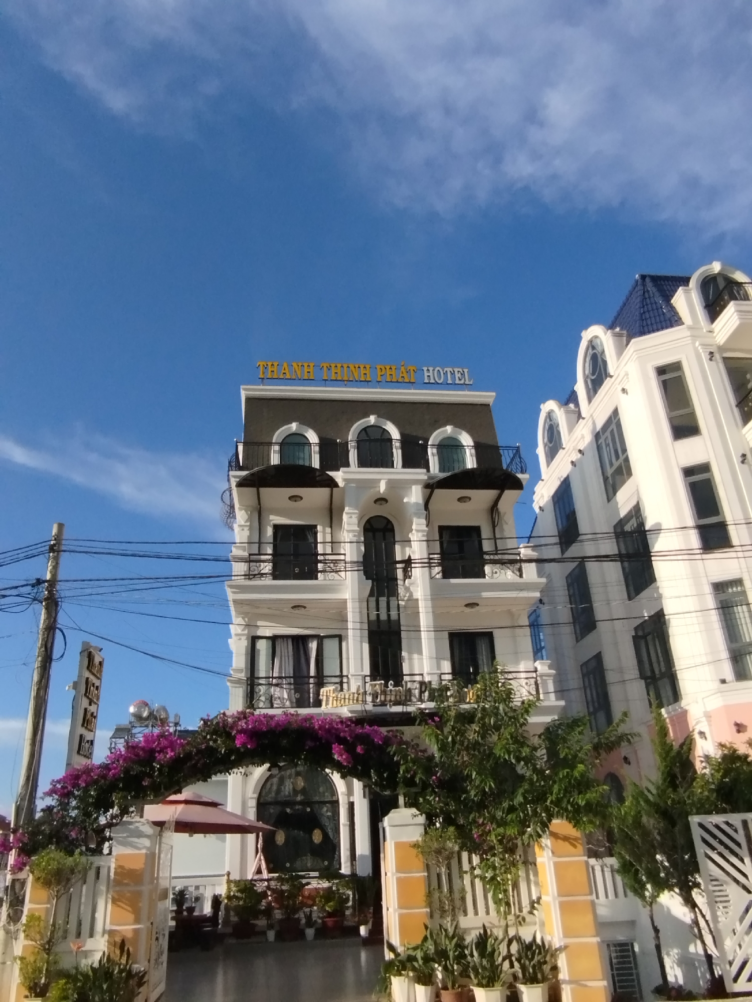 The image shows the Thanh Thinh Phat Hotel in Da Lat, Vietnam. The hotel is a multi-story building with a white facade, black trim, and a dark gray roof. A large yellow sign on the top of the building reads "THÀNH THỊNH PHÁT HOTEL". In front of the hotel, there is an archway covered in purple flowers, and a red umbrella. The sky is blue with some white clouds. There are power lines and a utility pole on the left side of the image. The architecture is reminiscent of colonial style buildings often found in Da Lat.