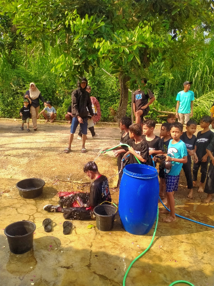 An outdoor scene in Gebyog, Indonesia, features approximately 15-20 individuals engaged in a water-dousing activity under bright sunlight. In the foreground, a young male wearing a black long-sleeved shirt and dark trousers sits on a wet, dirt-covered concrete surface, being actively sprayed with water from at least two green hoses held by young boys. Red and black debris, possibly confetti or mud, adheres to his clothing and the surrounding wet ground.
To the right of the seated male, a large blue plastic barrel stands upright, with a green hose visible near its base. Several boys, mostly clad in dark t-shirts, surround the seated male; two are actively spraying water, while others observe. One boy wears a light blue t-shirt and patterned shorts, holding a black bucket.
On the ground to the left, two black plastic tubs and a pair of black flip-flops are positioned on the wet concrete. In the mid-ground, a young male in a black hooded jacket, dark shorts, and sandals stands observing the central activity. To his left, an adult female in a light brown hijab and light pants walks, holding the hand of a small child in a black t-shirt. Another male in a light blue t-shirt and light pants stands further right in the mid-ground.
The background consists of dense, verdant foliage, including trees and tall grasses, under clear daytime conditions. Shadows are visible, indicating strong sunlight. The ground transitions from wet concrete in the foreground to packed earth or dirt further back.
