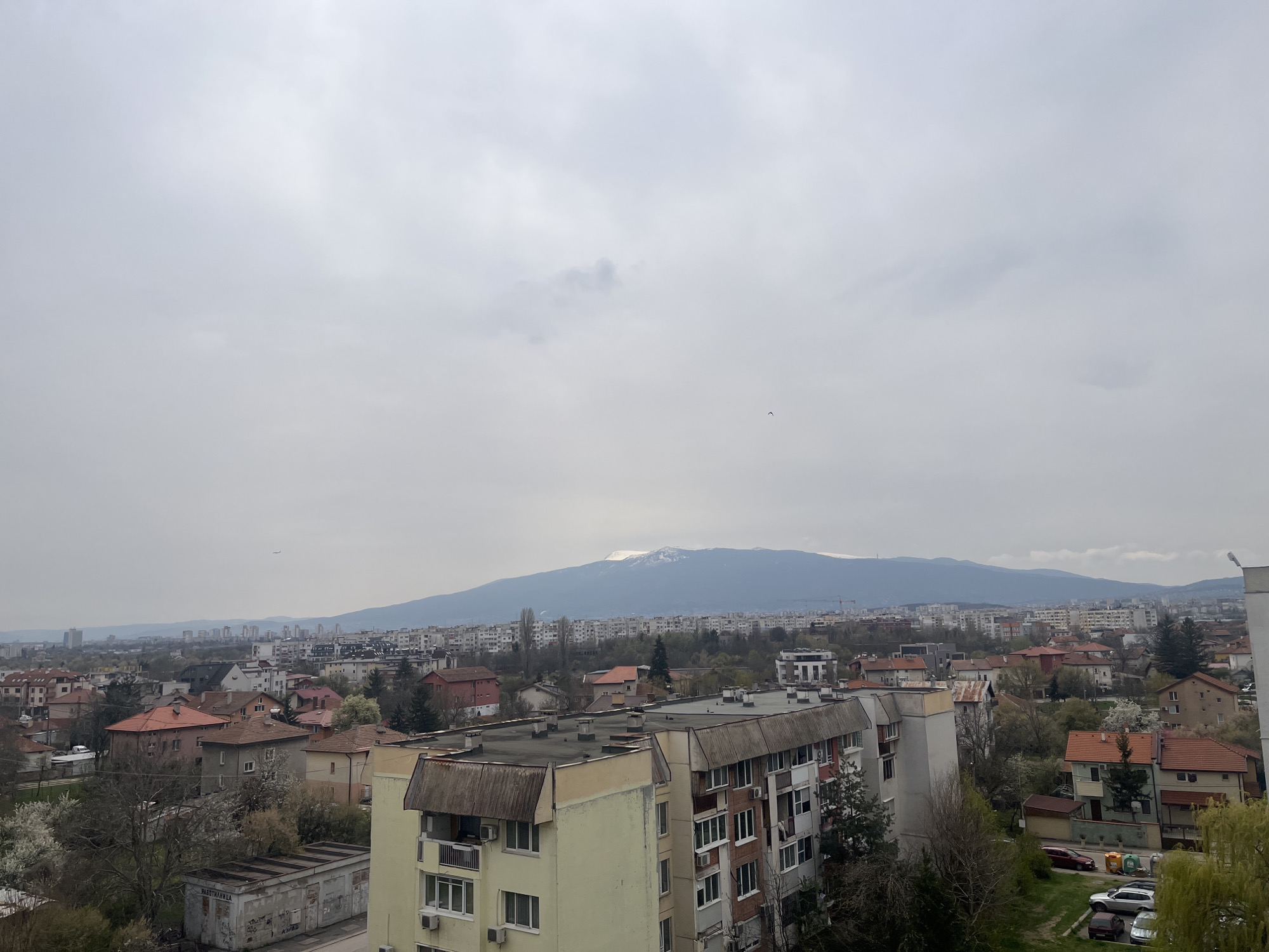 The image shows a panoramic view of a residential area in Sofia, Bulgaria, under an overcast sky. In the foreground are several multi-story apartment buildings with visible windows, balconies, and roofing details. The architecture appears typical of Soviet-era construction. Beyond these buildings stretches a cityscape of houses and more apartment blocks interspersed with trees. In the distance, a mountain range is visible, with traces of snow on the higher elevations. The overall environment suggests an urban setting on a cloudy day.