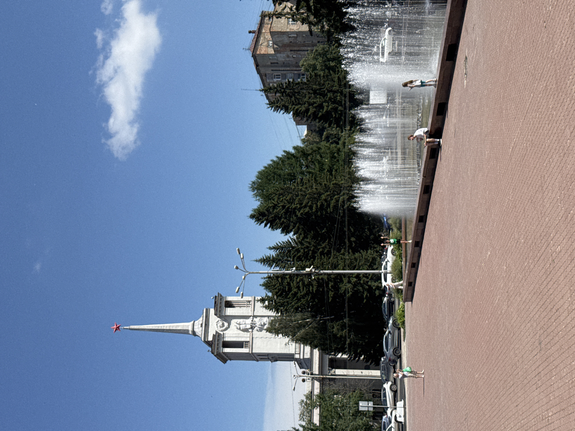 This is a vertically oriented, wide shot of a public square in Yekaterinburg, Russia. A prominent, white stone building with a red star on its spire dominates the left side of the frame. The building has architectural details and appears to be a historical monument or significant structure. In the background, a fountain with multiple streams of water is visible, with several people enjoying the spray. The ground is paved with red brick. The time of day appears to be daytime with a clear blue sky and a single, wispy white cloud. A line of cars is parked along the side of the square, suggesting an urban environment. One person is seen bending over near the fountain, and another is standing nearby. A couple of individuals are visible in the background by the fountain. The overall atmosphere seems peaceful and leisurely, with people enjoying a sunny day in a public space. There is no visible text in the image.