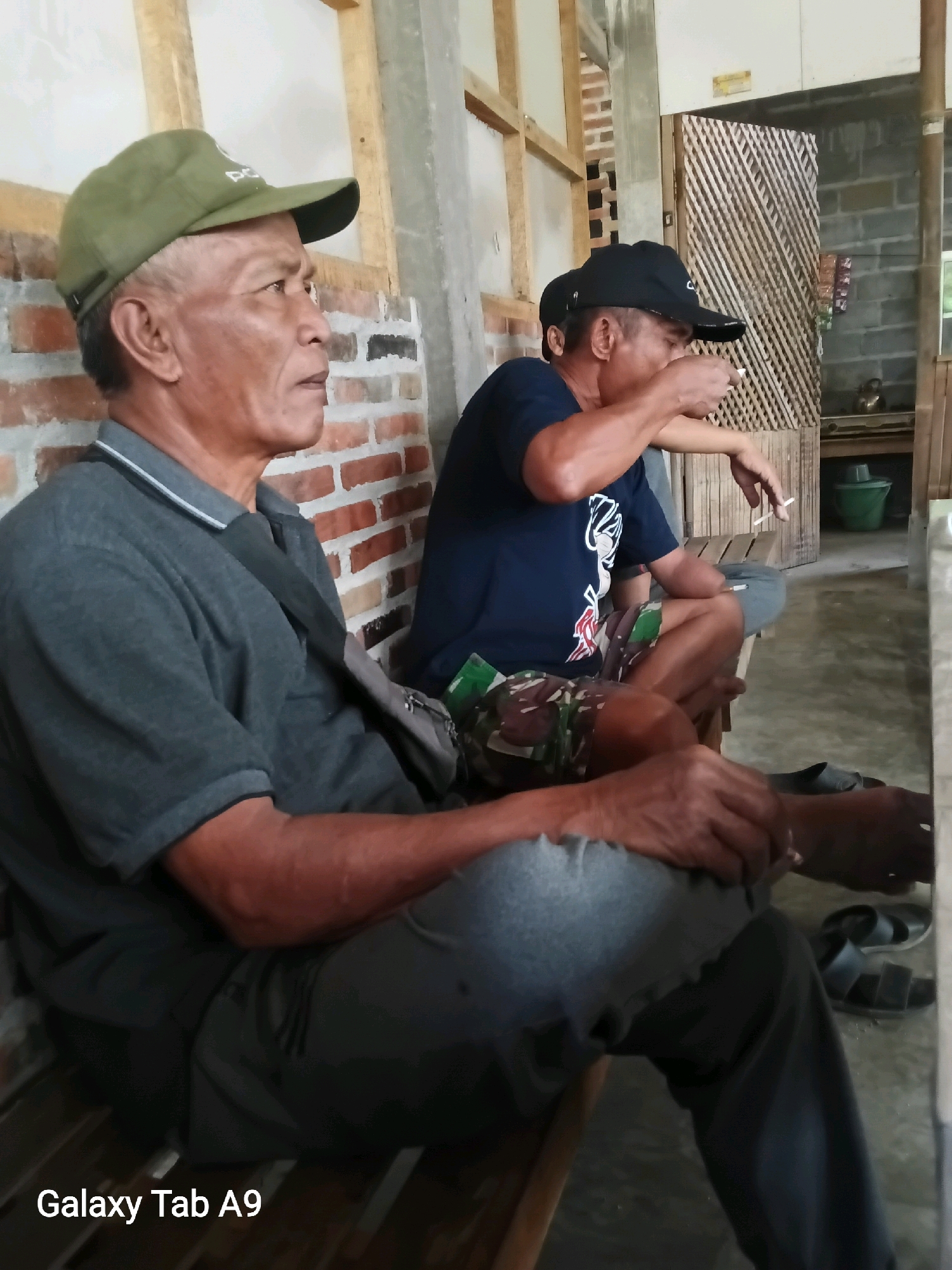 Two middle-aged Indonesian men are seated on a wooden bench, likely in a communal or outdoor setting in Ngadirenggo, Indonesia. The man on the left, wearing a green cap and a grey polo shirt, is looking to his right. The man on the right, wearing a black cap and a dark blue t-shirt with camouflage shorts, is holding a cigarette to his lips. Both men appear relaxed. The background features a brick wall, wooden beams, and a woven bamboo partition, suggesting a rustic or traditional building. Several pairs of sandals are visible on the floor. A watermark in the lower left corner indicates the image was taken with a "Galaxy Tab A9".