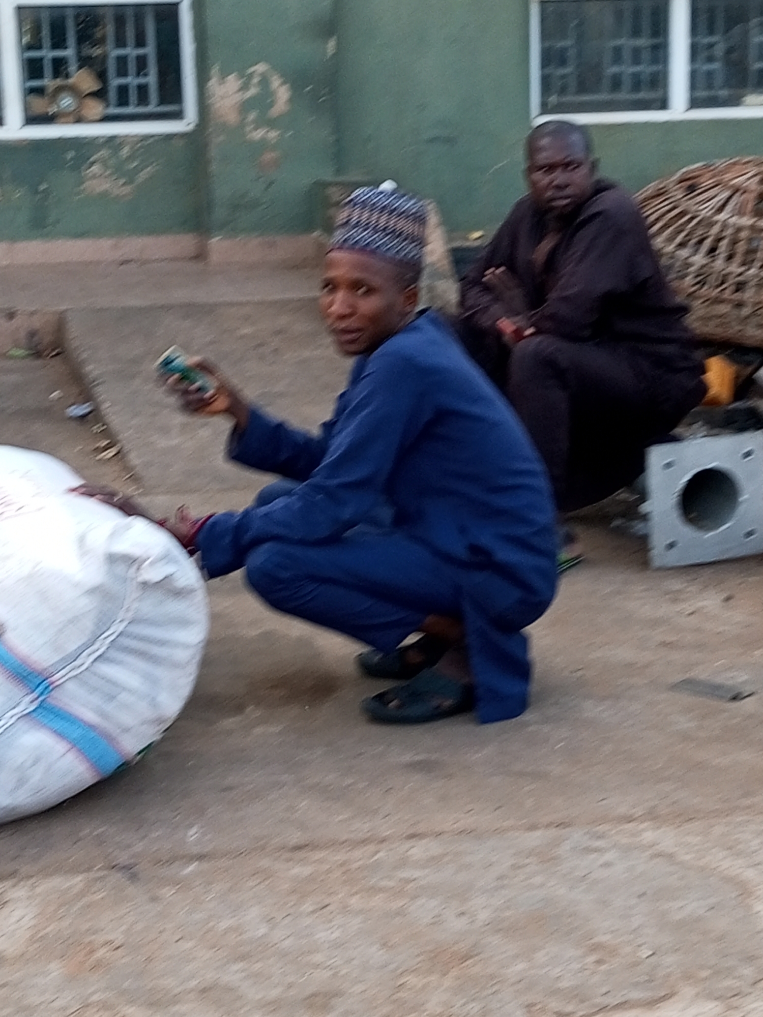 In the city of Gombe, Nigeria, two men are present in an outdoor area. The man in the foreground is squatting and wearing a blue garment and a patterned cap. He is holding a small green cylindrical object in his right hand and looking towards the camera. To his left, a large white sack is visible. In the background, a second man is seated, wearing a dark outfit and with his arms crossed. He appears to be facing slightly away from the camera. Behind him is a woven structure and a metallic object with a circular opening and bolt holes. The environment features a wall with two windows in the background, painted a green-grey color, and a paved or concrete ground surface.