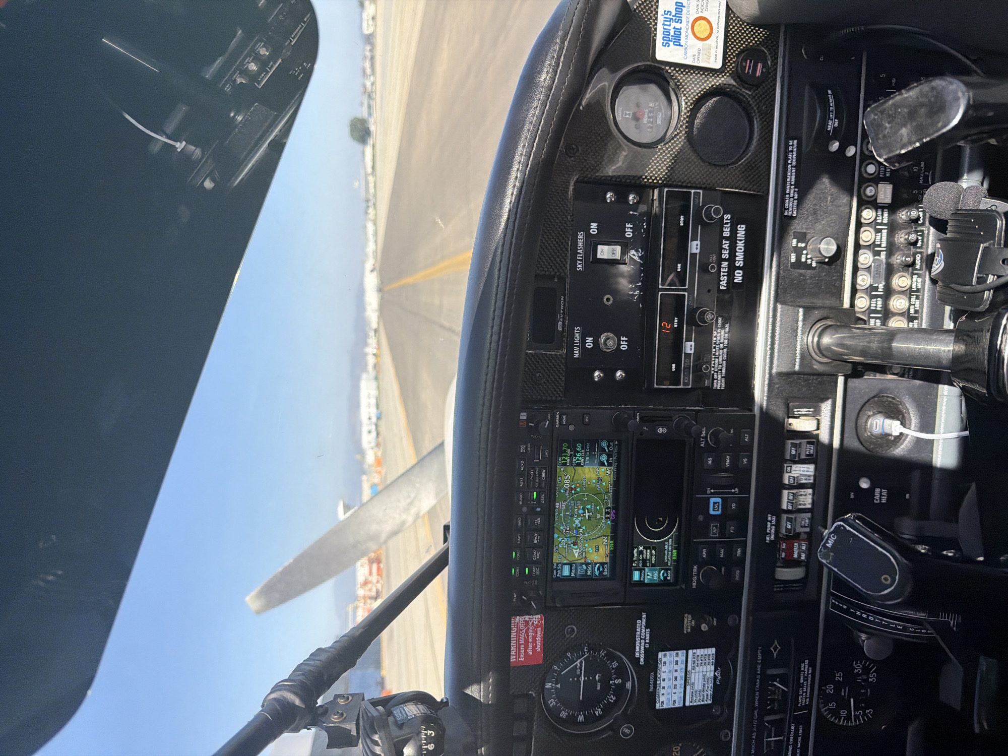 The image shows the cockpit of a small airplane, viewed from the pilot's perspective, preparing for takeoff from a runway. The plane's instrument panel is prominent, displaying various gauges, switches, and navigation equipment, including a Garmin GPS unit. Outside the aircraft, a runway stretches out towards the horizon, with buildings visible along the edge of the airfield. The aircraft is likely in the city of Los Angeles, United States of America.