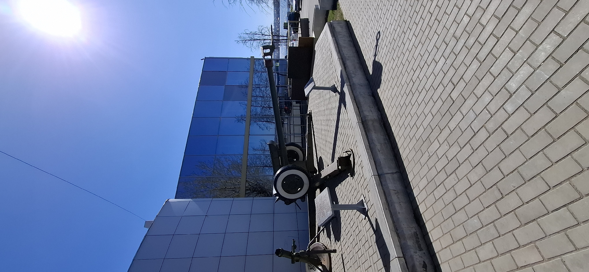 The image shows a modern building with a blue glass facade and a light gray brick sidewalk. A historical artillery piece is mounted on the sidewalk near the building. The location is Votkinsk, Russia, identified by the artillery monument, a memorial to the city's contribution to the arms industry. The sky is clear blue, and the sun is visible in the upper left.