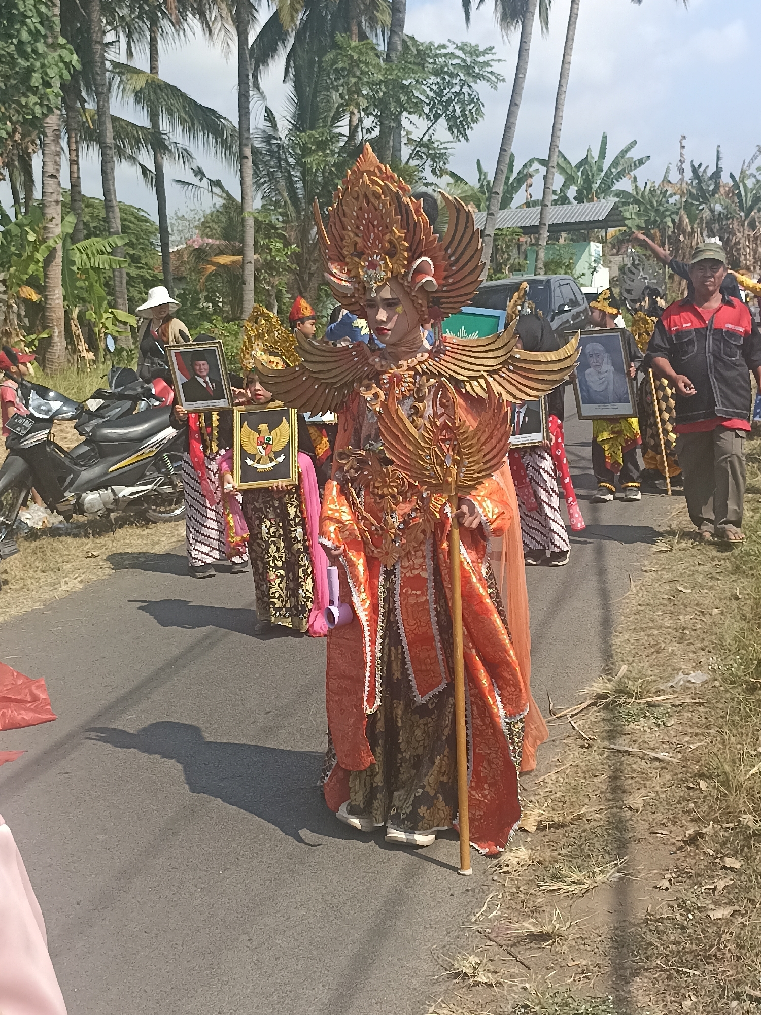 A young person in elaborate, golden, and orange ceremonial costume with a large headdress walks down a paved road in Banyuwangi, Indonesia. The costume features intricate wing-like adornments and a long, flowing skirt. The person holds a wooden staff. Behind them, other participants in traditional attire are visible, some carrying framed portraits and a framed emblem of the Garuda Pancasila, the national symbol of Indonesia. A black motorcycle is parked on the left side of the road. The background shows a line of tall palm trees and dense vegetation. The sky is overcast. The scene appears to be a parade or cultural event.