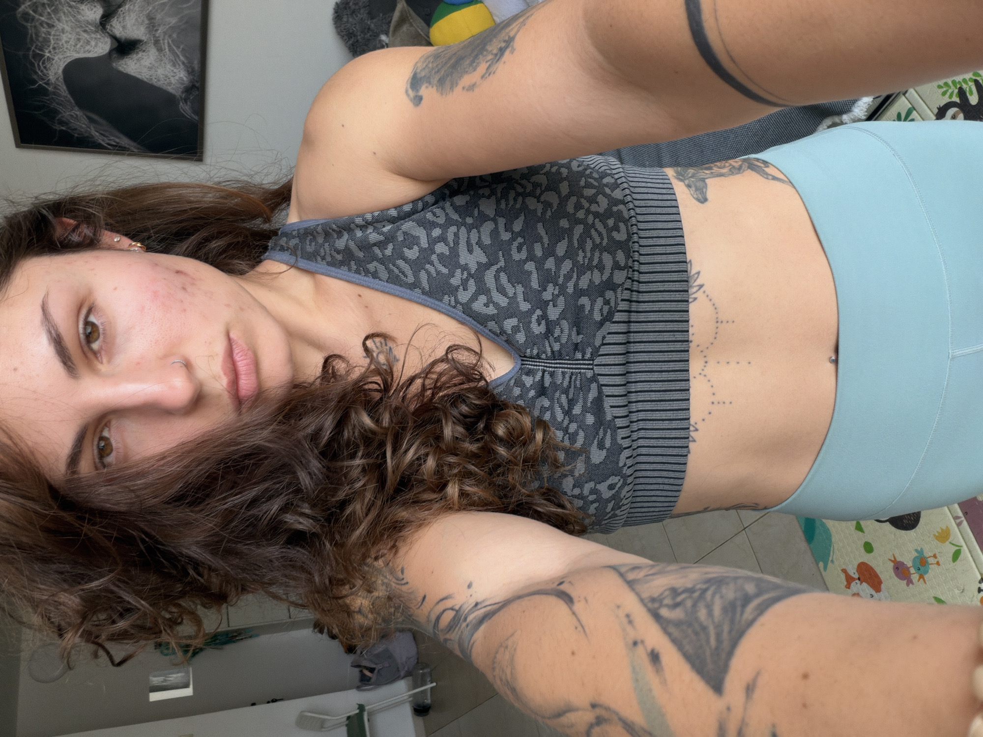 A young adult woman with brown curly hair, acne on her face, and multiple tattoos is posing for a selfie. She is wearing a gray animal print sports bra and light blue leggings. The background includes a framed picture on the wall, a playful mat with cartoon animals, and other household objects. While the city is mentioned, there is no information available from the image itself to corroborate or confirm that this image was taken in Kiryat Bialik, Israel.