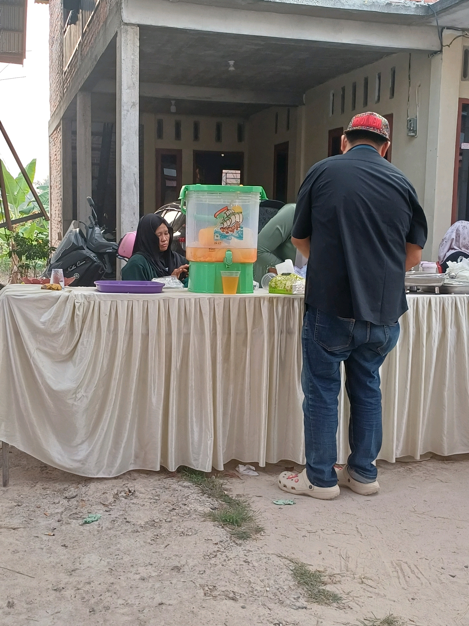 In Dumai City, Indonesia, a scene unfolds under an elevated building structure. A table draped with a cream-colored cloth dominates the foreground, set up for an outdoor event. On the table, a large, clear plastic beverage dispenser with a green lid holds a yellow liquid, likely juice. Beside it, a woman wearing a black hijab and green top is seated, appearing to be serving or managing the drinks. A purple tray sits in front of her, and a glass of the yellow liquid is visible to her right. To the left, a motorcycle is parked.
A man stands facing away from the camera, toward the table, dressed in a dark short-sleeved shirt, blue jeans, and casual clogs. He wears a red and white patterned baseball cap. Another person, partially visible and wearing a light-colored headscarf, is seated at the far right of the table, behind a serving tray. The ground is sandy and littered with small debris. The background shows a building with an open facade, suggesting it's a residence or a community space. The overall impression is that of a local gathering or a small-scale catering event.