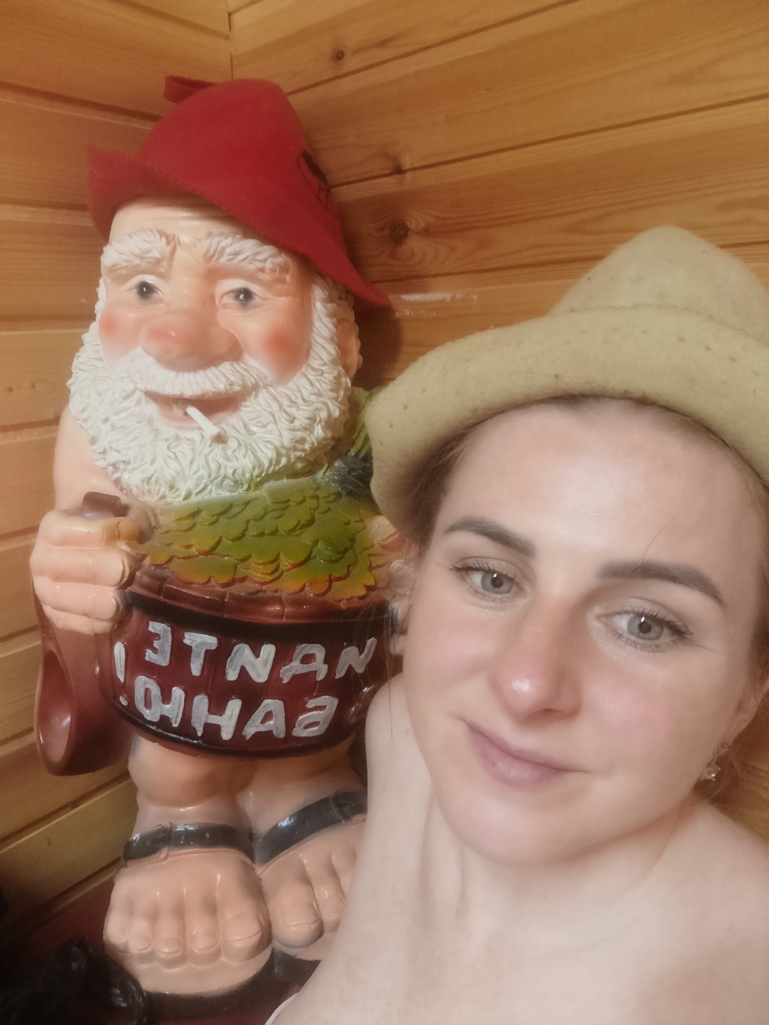 A person wearing a hat is in a sauna next to a gnome statue holding a beer mug that has "NANTE SAUNA" written on it. The sauna has light wood paneling.