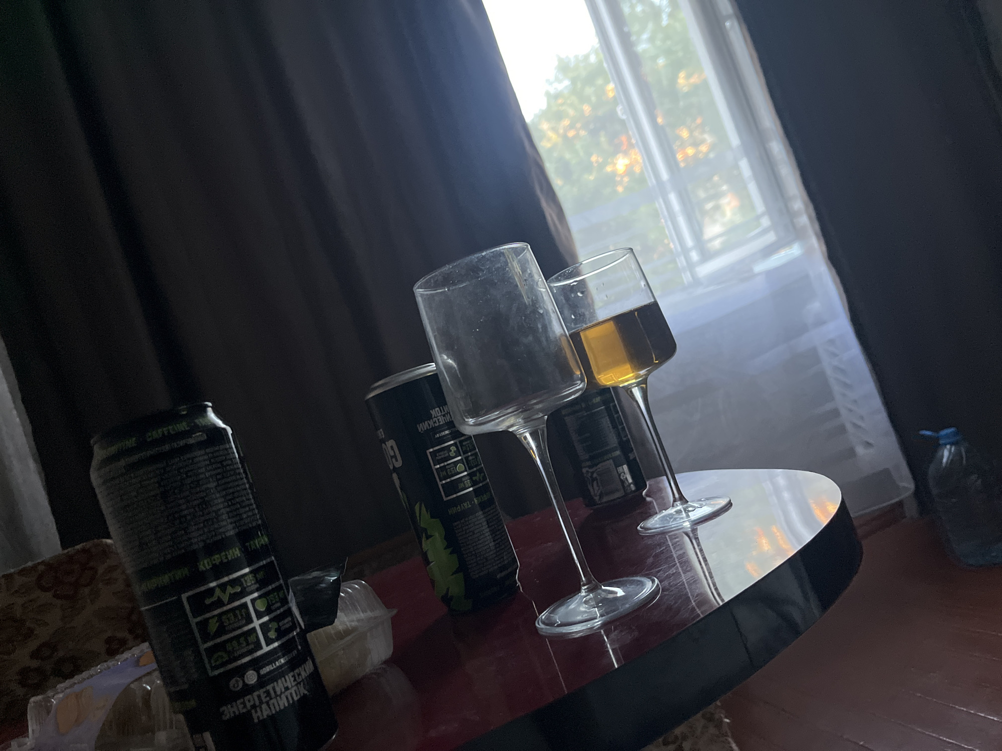 The image depicts a dark room with a table in the foreground. On the table, there are two wine glasses, one empty and the other containing a golden liquid. Two energy drink cans are also visible, one prominently displaying Russian text that translates to "Energy Drink." The room is dimly lit, with natural light filtering through a window covered by a sheer curtain, suggesting it may be daytime. The overall atmosphere is casual, possibly indicating a domestic setting. The location in Svietlahorsk, Belarus, is not directly discernible from the image's visual elements but is provided as context.