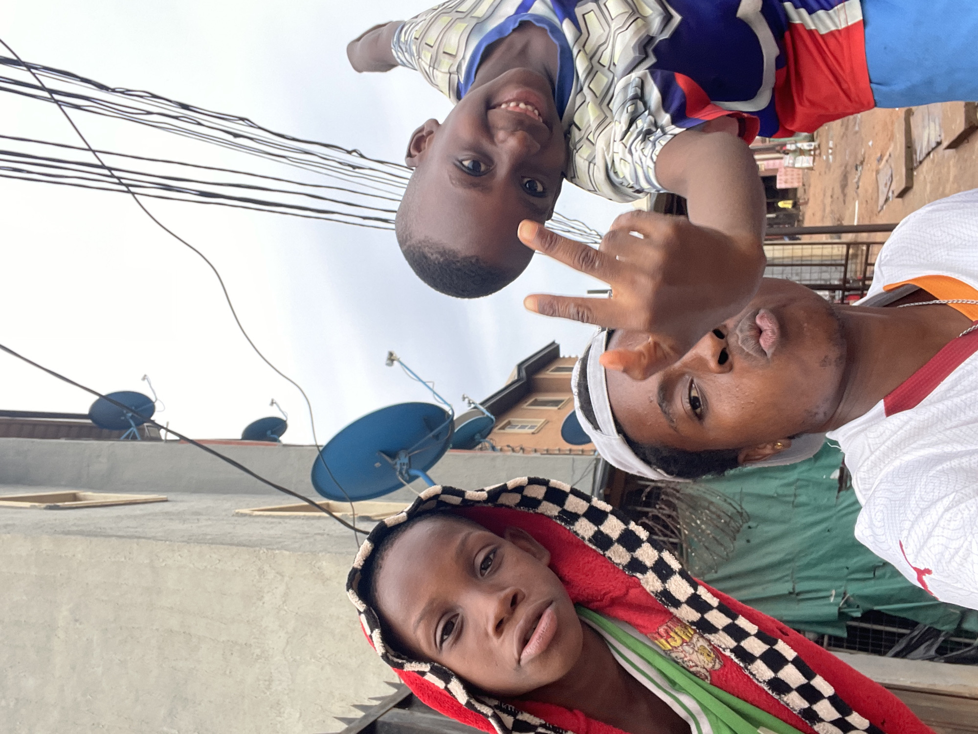 Three dark-skinned individuals are pictured in Onitsha, Nigeria. A child wearing a patterned shirt and the peace sign is at the top, followed by a man in a white shirt. The third person, in the foreground, is wearing a hooded, red and checkered scarf. Satellite dishes are visible atop nearby buildings with electrical wires running across the sky. A weathered building forms the backdrop.