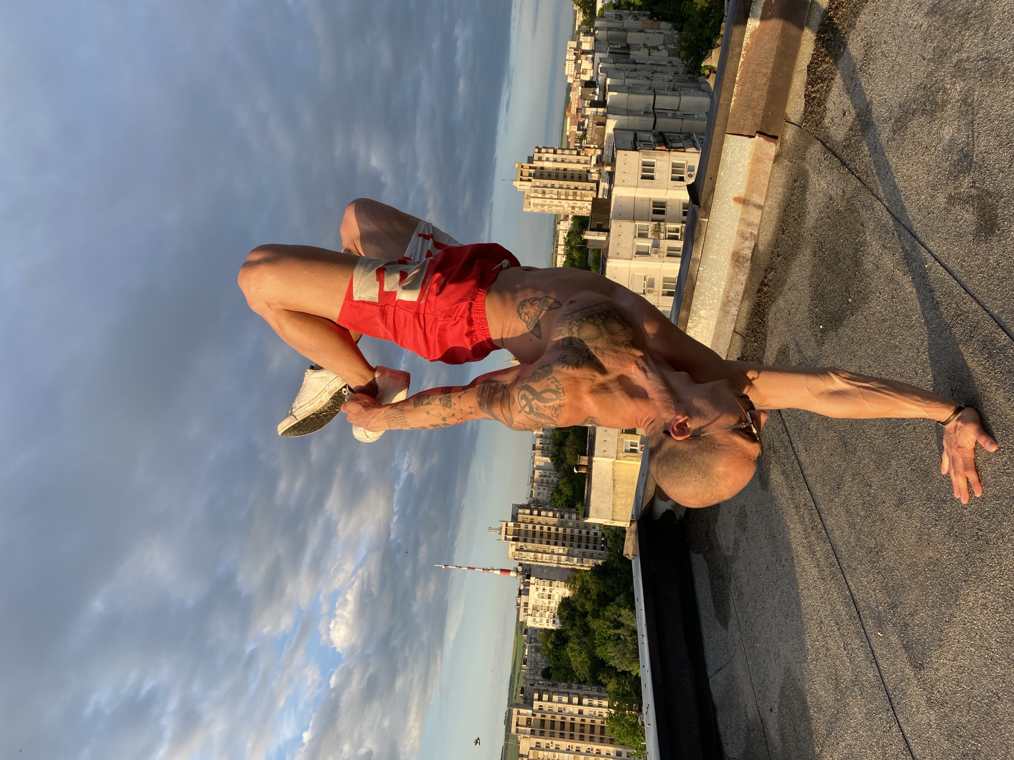 A highly flexible, muscular man with numerous tattoos is captured mid-pose on a building rooftop, performing a deep backbend. He is shirtless, wearing red shorts with white accents, white sneakers, and sunglasses. His bald or closely-shaved head is tilted back, looking towards the cloudy sky. One hand is planted on the rough, dark rooftop surface for balance, while the other arm is extended. His legs are bent, with his feet lifted high towards his head.
The setting is a building rooftop, characterized by its dark, coarse surface and a rusty metal coping along the edge, with a thin cable also visible. In the background, a sprawling cityscape unfolds under a partly cloudy sky illuminated by the golden light of either sunrise or sunset. Numerous multi-story residential buildings, typical of post-communist architecture in Pleven, Bulgaria, extend into the distance. A prominent red and white telecommunications tower stands tall amidst the urban landscape. Patches of green trees are visible between the buildings. The overall scene suggests a moment of physical prowess and tranquility above the city. No discernible text is visible.