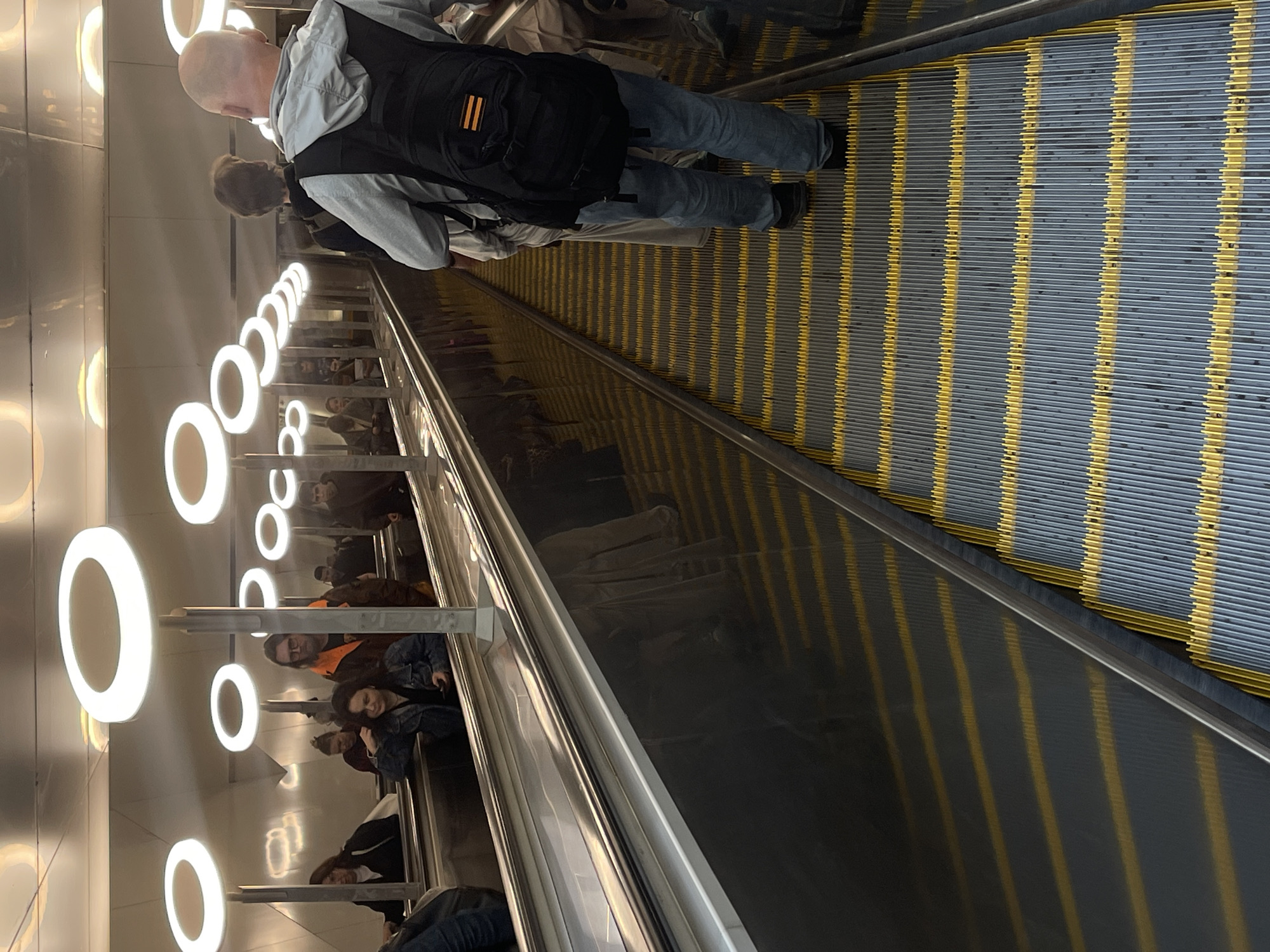 The image depicts an escalator in an underground station, filled with people riding in both directions. The escalator has a metallic gray and yellow striped surface and a silver handrail. A bald man wearing a gray jacket, blue jeans, black shoes, and a black backpack is riding up the escalator, facing the viewer. Many other people of various ages and clothing are also on the escalator. The walls of the station are white with a row of circular white lights above the escalator. The image is taken from a slightly elevated perspective. The location is Unknown, Unknown.