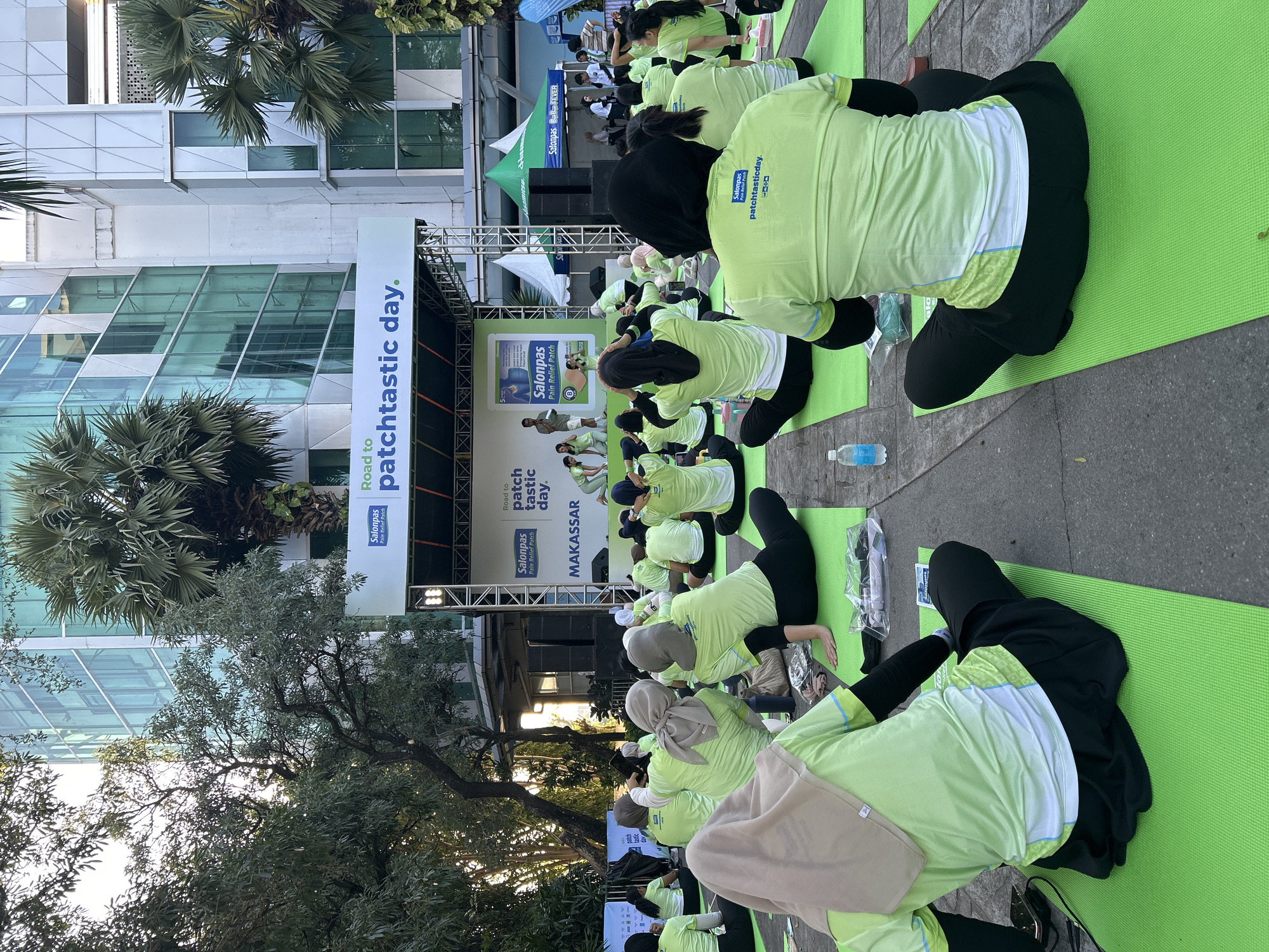 In Makassar, Indonesia, a group of women are participating in an outdoor yoga session on a paved area in front of a modern, glass-fronted building. They are seated on bright green yoga mats, mostly wearing light green t-shirts and black bottoms. Some women are wearing headscarves in various colors like beige and gray. In the background, a stage is set up with a banner that reads "Road to patchtastic day." and "Salonpas". A few individuals are standing on the stage, seemingly leading the session or giving instructions. Trees and palm trees surround the area, and sunlight filters through the leaves, indicating it is daytime. Several water bottles are visible on the mats.