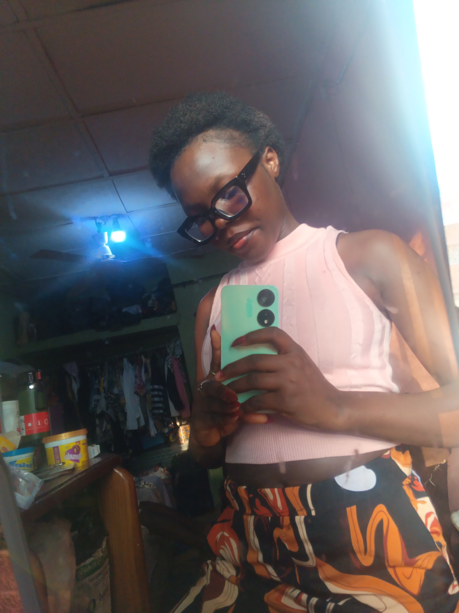 A young Black woman is taking a mirror selfie in what appears to be a bedroom or dressing area in Onitsha, Nigeria. She is wearing a pale pink sleeveless top with a ribbed texture and a pair of patterned trousers with a black background and orange, white, and brown wavy designs. She has on large, thick-rimmed black glasses and is holding a mint green smartphone in her right hand. The background is cluttered, with a closet filled with clothes visible behind her, and shelves with various items, including containers and bottles, are to her left. A ceiling fan is mounted on the ceiling, and a bright blue light fixture is also visible. The overall lighting suggests it might be daytime, but the indoor setting and reflections indicate a controlled environment.
