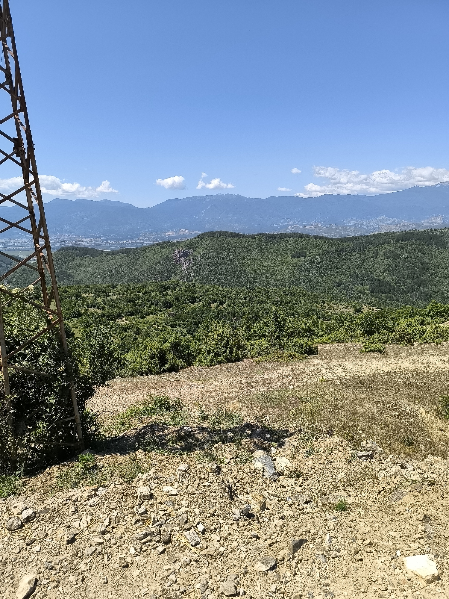 The image captures a panoramic view of the mountainous terrain surrounding Gorno Dryanovo, Bulgaria, under a clear blue sky with scattered white clouds. In the foreground, a rusty metal pylon, part of an electricity transmission tower, dominates the left side of the frame. Its latticework structure is weathered and shows signs of age. The ground in the immediate foreground is rough and rocky, with patches of dry grass and soil.
Beyond the pylon and foreground, the landscape descends into a lush, verdant valley. Dense green forests cover the rolling hills, interspersed with smaller trees and shrubs. The vegetation appears healthy and vibrant, suggesting a temperate climate.
In the mid-ground, a saddle between two hills reveals a glimpse of a town or village nestled in the distance, with buildings and infrastructure faintly visible. The middle and background are characterized by layers of mountains, progressively fading into a hazy blue as they recede into the distance. The overall scene conveys a sense of natural beauty and remoteness, with the man-made structure of the pylon providing a contrasting element to the wild landscape.