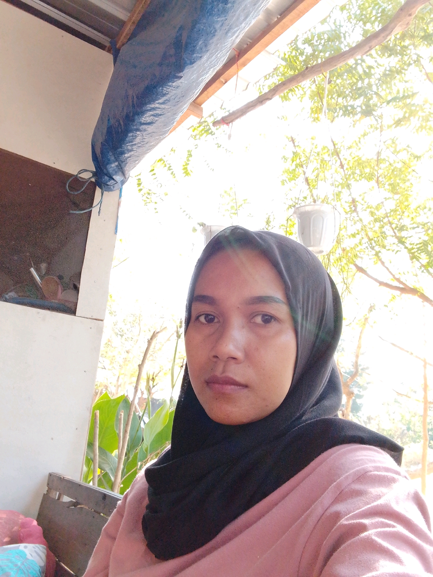 A medium shot photograph captures a woman wearing a black hijab and a pink shirt, facing the camera. She is positioned in front of a white wall and a wooden structure, with lush green plants and trees visible in the background, illuminated by bright sunlight. The scene appears to be outdoors, possibly in a residential area or a garden. The woman's expression is neutral, and she is the central focus of the image. There are no explicit indications of the city of Cikal, Indonesia, within the image itself, making it impossible to confirm the location.