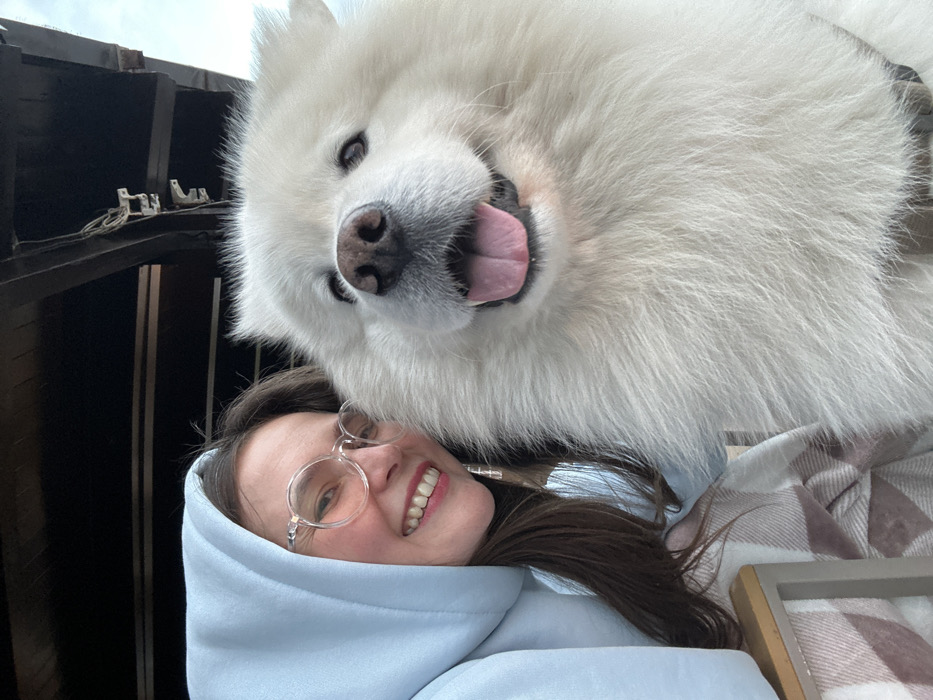 A close-up photograph captures a female subject and a large white Samoyed-type dog in the city of Šušanj, Montenegro. The female is positioned on the left, facing slightly right, wearing a light blue hooded sweatshirt with the hood up, and clear-framed round eyeglasses. She has visible dark hair and is smiling, revealing her teeth. Her left forearm rests on a surface covered by a light-colored plaid blanket. To the right, the white-furred dog faces the camera with its mouth open and tongue partially extended. A dark harness or collar is visible on the dog's lower neck/chest area. The background features dark wooden beams and planks, consistent with an overhead structure like a patio roof, with a brighter sky visible above. A thin wire with small white clips is attached to one of the wooden beams.