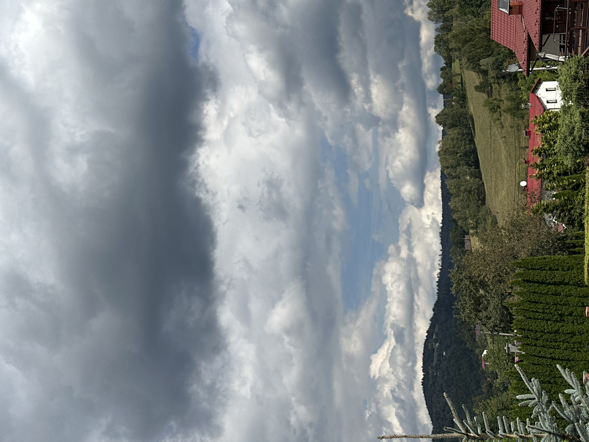 This image captures a panoramic view of a rural landscape in Wisła, Poland, under a dramatic sky. The foreground is dominated by a tall, blue-green conifer tree, its branches reaching towards the viewer. Immediately behind it, a dense, dark green hedge forms a sharp horizontal line across the frame.
Beyond the hedge, the scene unfolds into rolling hills covered in lush green vegetation and scattered trees. Several houses with red-tiled roofs are visible, nestled among the trees and on the slopes of the hills. One house, painted white with red trim, stands out in the mid-ground. Further in the distance, a darker, tree-covered mountain range forms the backdrop, its silhouette softened by the atmospheric conditions.
The sky is a prominent feature, filled with a complex arrangement of clouds. Large, dark grey storm clouds dominate the upper portion of the frame, suggesting an impending or passing rain shower. Lighter, white clouds drift below, allowing patches of pale blue sky to peek through in the middle distance. The lighting is diffuse, casting a subdued light over the entire scene, with some areas of the landscape appearing brighter where the clouds are thinner.
No people or specific actions are depicted in this static landscape view. The environment is natural and rural, characterized by hills, forests, and residential structures.
