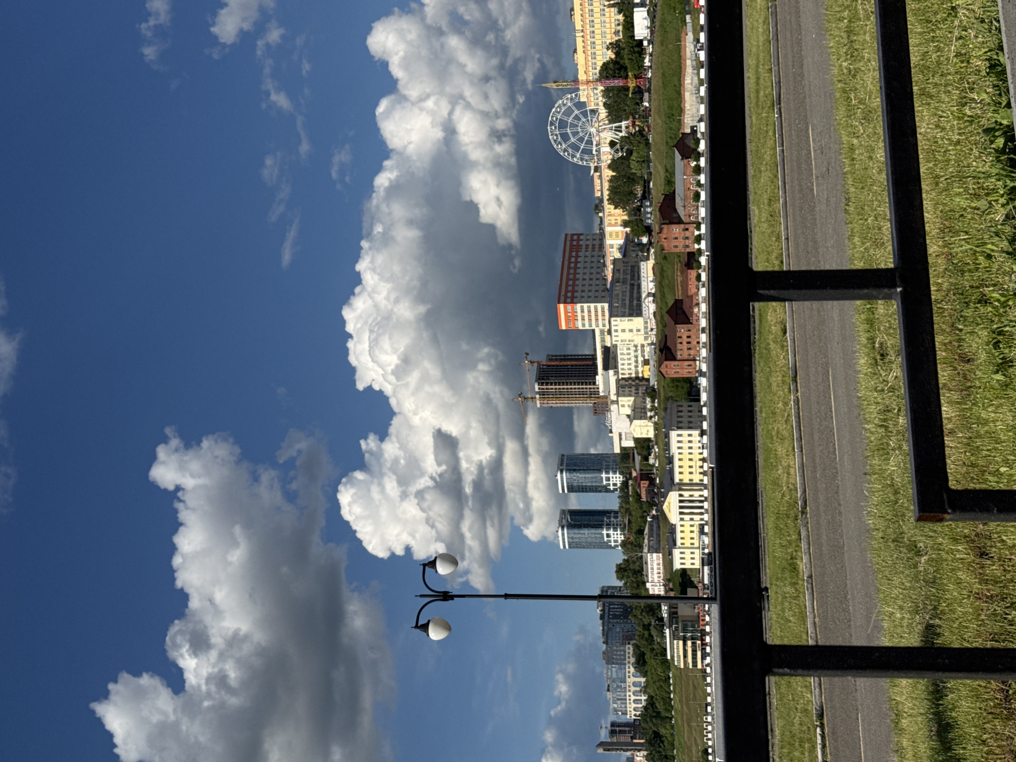 The image shows a view of the city of Izhevsk, Russia, looking towards the city center from a slightly elevated position, likely a park or pedestrian area, as evidenced by the presence of a black metal fence in the foreground and a paved pathway to the right. The sky is blue with large, white cumulus clouds. In the distance, a Ferris wheel is visible, a known landmark of Izhevsk Central Square. Several high-rise buildings are scattered throughout the cityscape, along with what appears to be an active construction site with a crane. The city's landscape includes a mixture of residential and commercial buildings, some of which have distinct architectural styles. A vintage-style streetlight with two globes is present in the middle ground, adding to the scene's character. The image is taken from a slightly tilted angle, offering a dynamic perspective of the city's panorama.