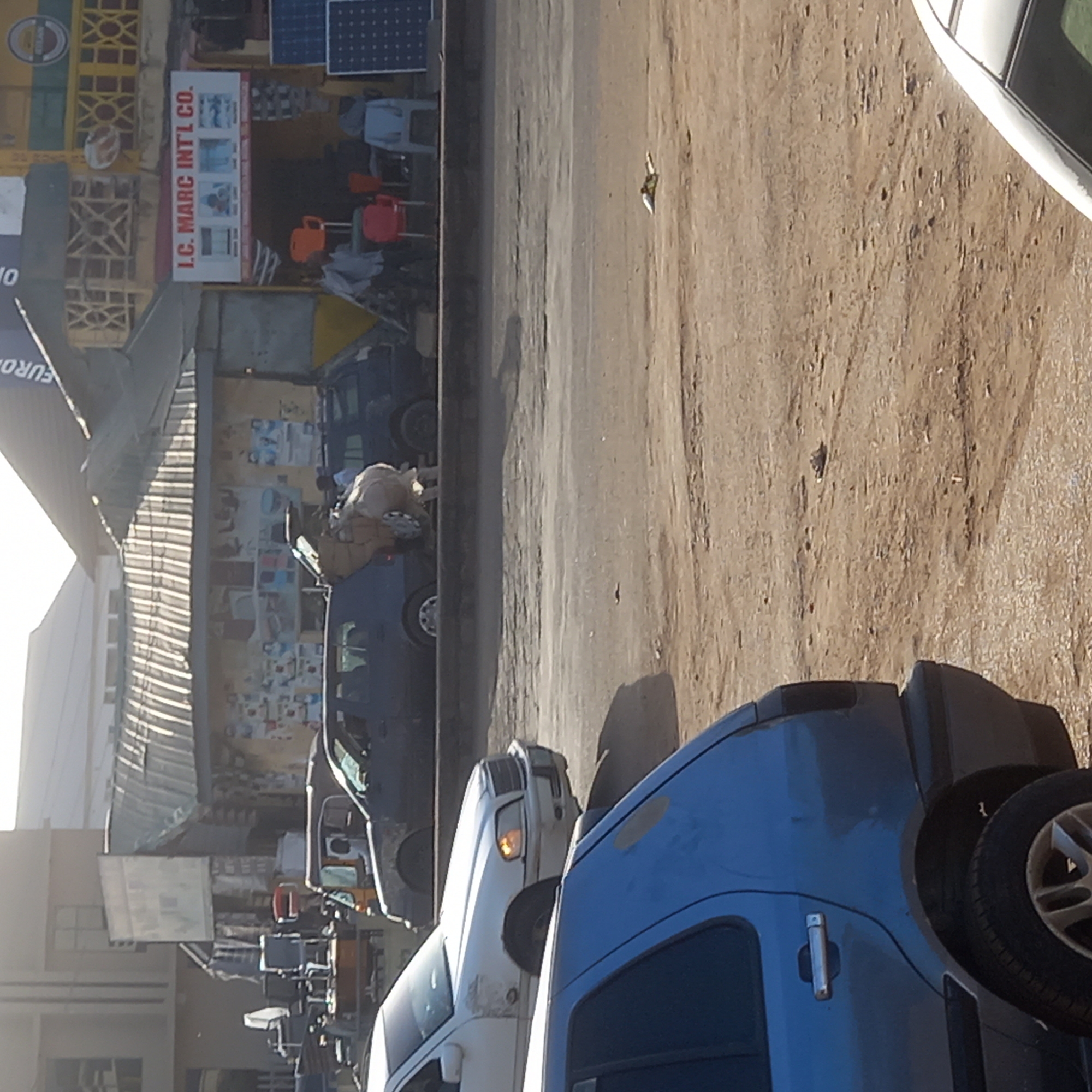 This image depicts a bustling street scene in Dadi Riba, Nigeria, under bright daylight. The ground is a dusty, unpaved road, showing signs of vehicle traffic.
In the foreground, a blue SUV with a rear-mounted spare tire is visible. Moving towards the background, a white pickup truck with its headlights on is present, followed by other dark-colored vehicles. Further down the street, a dark blue car is being loaded or unloaded by a person, suggesting commercial activity or transportation of goods.
Along the roadside, several shops or stalls line the street. A notable establishment displays a white sign with red text that reads, "I.C. MARC INT'L CO." Below this text, images suggesting home appliances or electronics are visible. Outside this shop, a variety of items are displayed, including plastic chairs in red, orange, and grey, and what appear to be large containers. On the roof above this shop, two solar panels are clearly visible. Another partially seen sign further down the street includes the word "EURO."
The strong shadows indicate a bright, clear day, likely morning or late afternoon. The scene captures a typical day in a commercial zone, highlighting local businesses, goods for sale, and the movement of people and vehicles in this Nigerian setting.