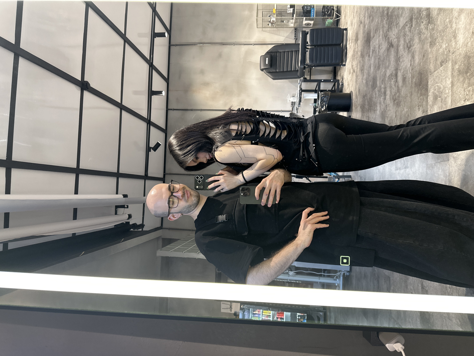 A man and a woman are reflected in a mirror, seemingly taking a selfie. The man has a shaved head, glasses, and a slight beard, wearing a black t-shirt and jeans. He holds a smartphone. The woman stands behind him, wearing a black top with cutouts in the back and black pants. She also holds a smartphone.
The reflection reveals a modern-looking interior. In the background, there is what appears to be barber chairs and equipment, suggesting a hair salon or barbershop. The walls are primarily white with black structural accents. There are rows of lights along the top of the mirror. Below, a sign for "Lager Beer" is visible, suggesting the image was taken at Lager Barbershop in Moscow, Russia.