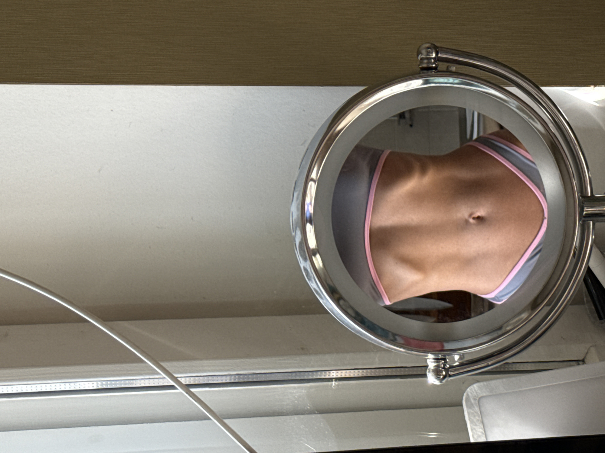 A close-up, slightly skewed, medium shot shows a round, chrome mirror reflecting a woman's abdomen, partially visible inside the frame. She is wearing grey and pink underwear. The mirror is on a white surface with a wooden panel in the background. The location cannot be determined.
