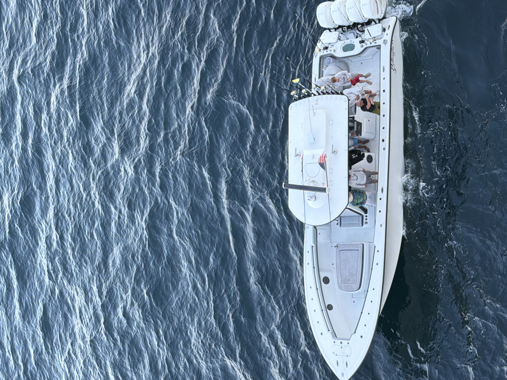The image shows a white boat moving on a dark blue body of water. The boat has multiple outboard motors at the stern and a group of people on board. The people are wearing casual clothing and appear to be relaxing. The boat is a center console type, and its name, "Yellowfin," is visible. An American flag is attached to a structure on the boat. The water has a textured surface, indicating wind or current. The image provides no location information to suggest a specific city or place.
