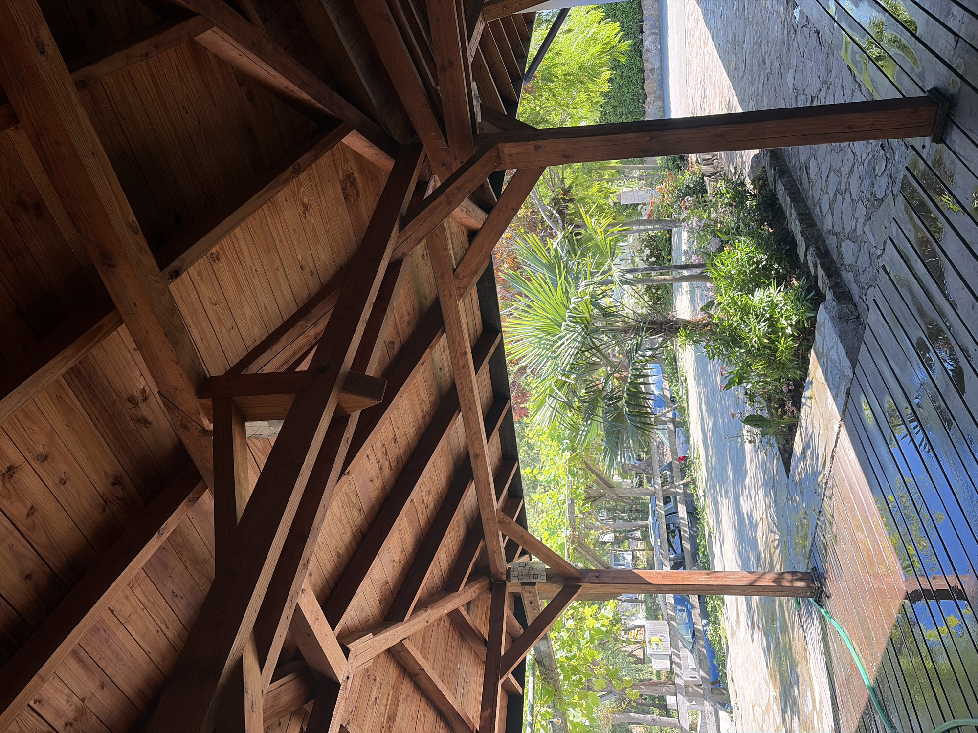 The image depicts the interior of a wooden gazebo with a complex, angular roof structure made of stained wood. Sunlight streams through the gaps in the roof, casting shadows and highlighting the texture of the wood grain.
Outside the gazebo, a pathway constructed of irregularly shaped stones leads away from the wooden deck. Lush green foliage and flowering plants are visible, with a palm tree standing prominently in the mid-ground. Beyond the garden area, a road is visible with parked cars. Trees and a stone wall line the background. The ground on the pathway and deck appears wet, suggesting recent watering or rain. A green hose is visible on the wooden deck, coiled near a wooden support pillar of the gazebo.
The scene is characteristic of an outdoor area in a warm climate, possibly a garden, resort, or a residential property. Given the context of Tsarevo, Bulgaria, this could be a tourist accommodation or a private residence near the Black Sea coast.