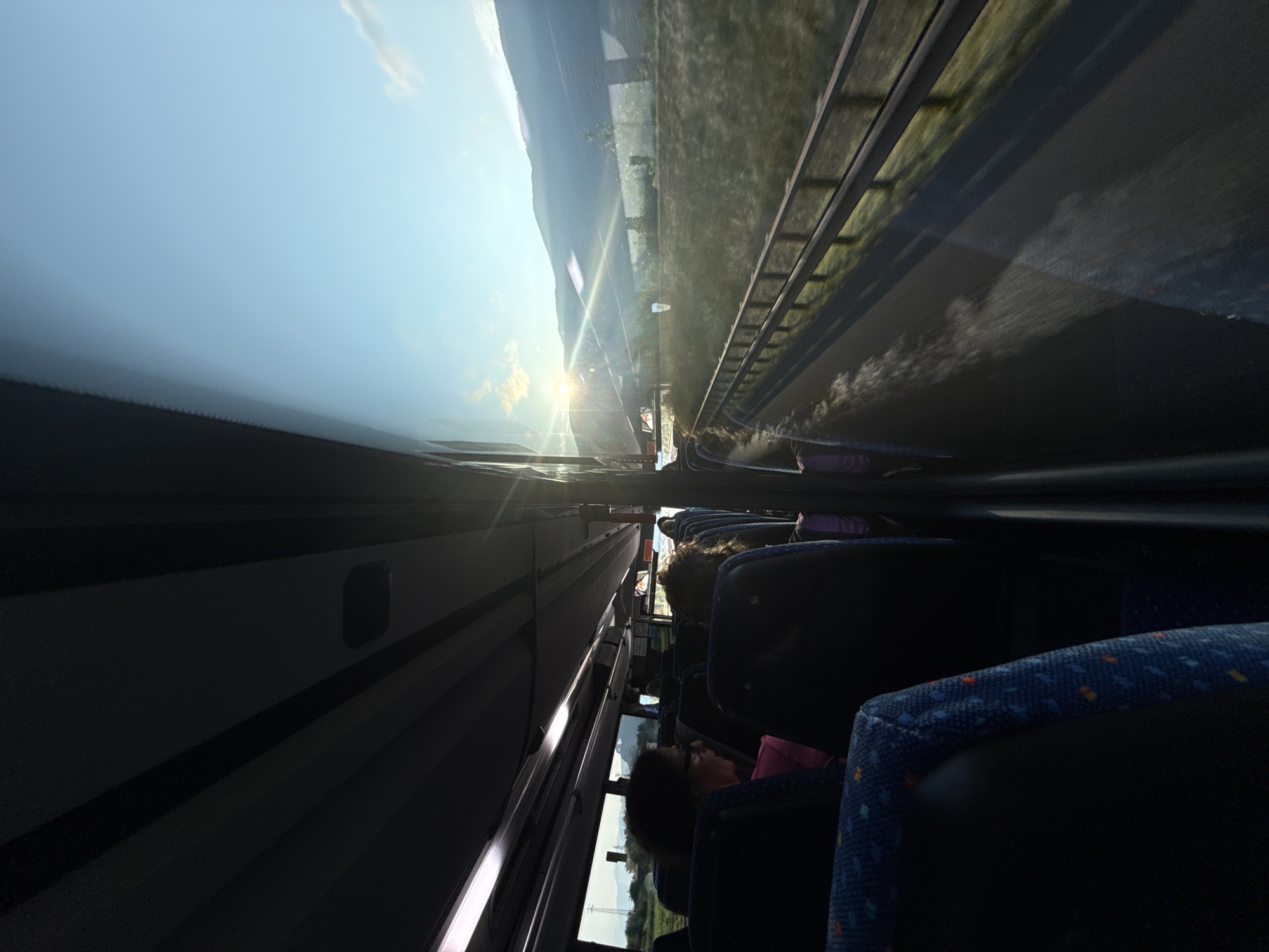 The image is an interior view from the passenger seats of a bus traveling along a highway. The sun is setting, casting a warm glow over the mountainous landscape visible through the windows. The bus has rows of dark seats with blue upholstery, and passengers are visible in some of the seats, looking out the window or facing forward. The road is a blur of motion, indicating the bus is in transit. The lighting suggests it is late afternoon or early evening. The scene is calm and relaxed, with a sense of travel and observation. There is no specific indication of the city of Neumarkt - Egna, Italy.