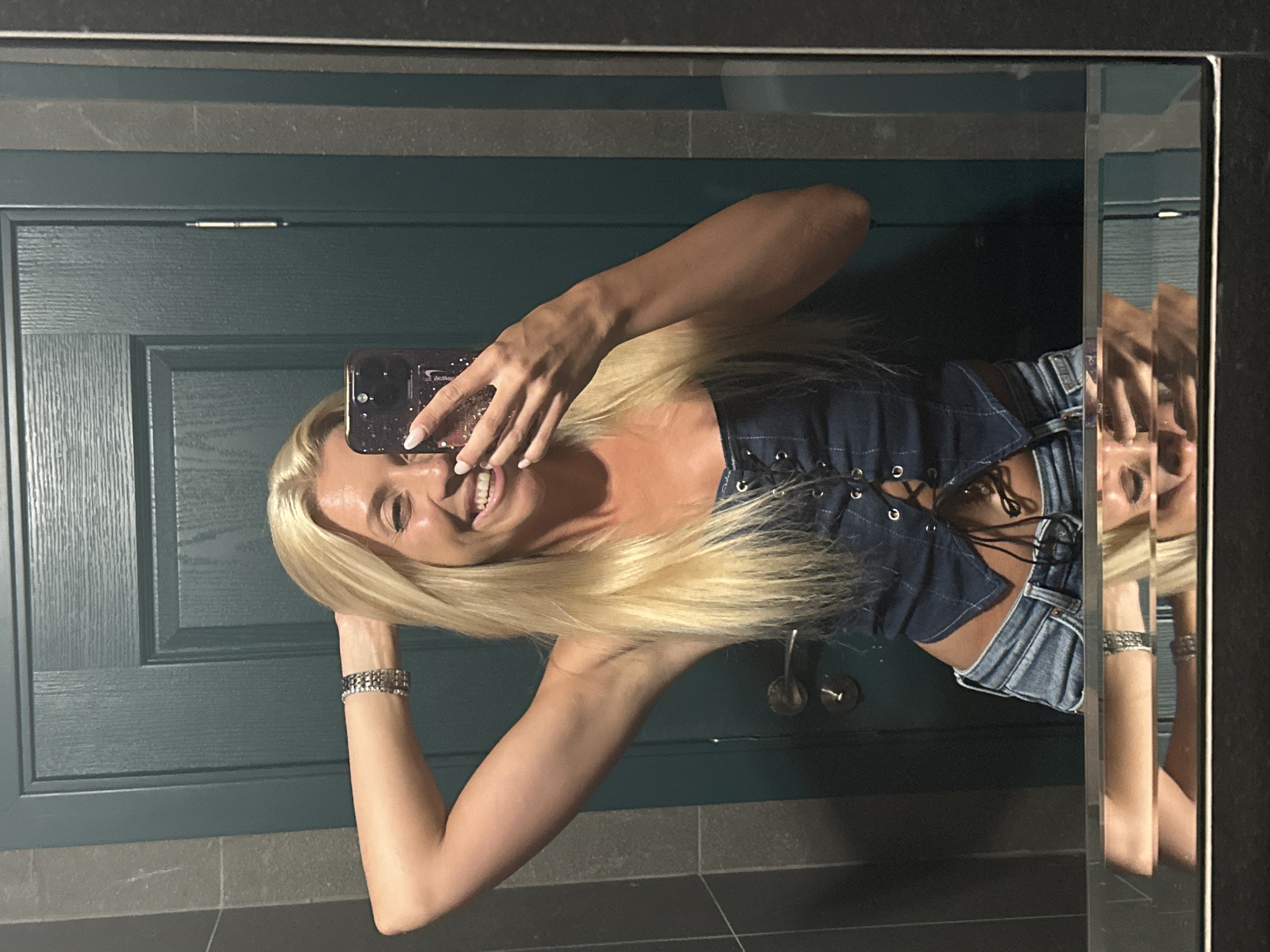 A woman with blonde hair and fair skin takes a mirror selfie in what appears to be a bathroom. She is wearing a dark blue denim corset-style top with lace-up detailing and light blue jeans. She is smiling broadly and has her right hand raised behind her head, with her left hand holding a smartphone to take the photo. A silver bracelet is visible on her left wrist. The background shows a dark teal door with metallic hardware and textured gray walls. The image is a reflection in a mirror, with the bottom of the frame showing a distorted reflection of the woman's lower body. The location in the city of Pazardzhik, Bulgaria, is not ascertainable from the provided image.