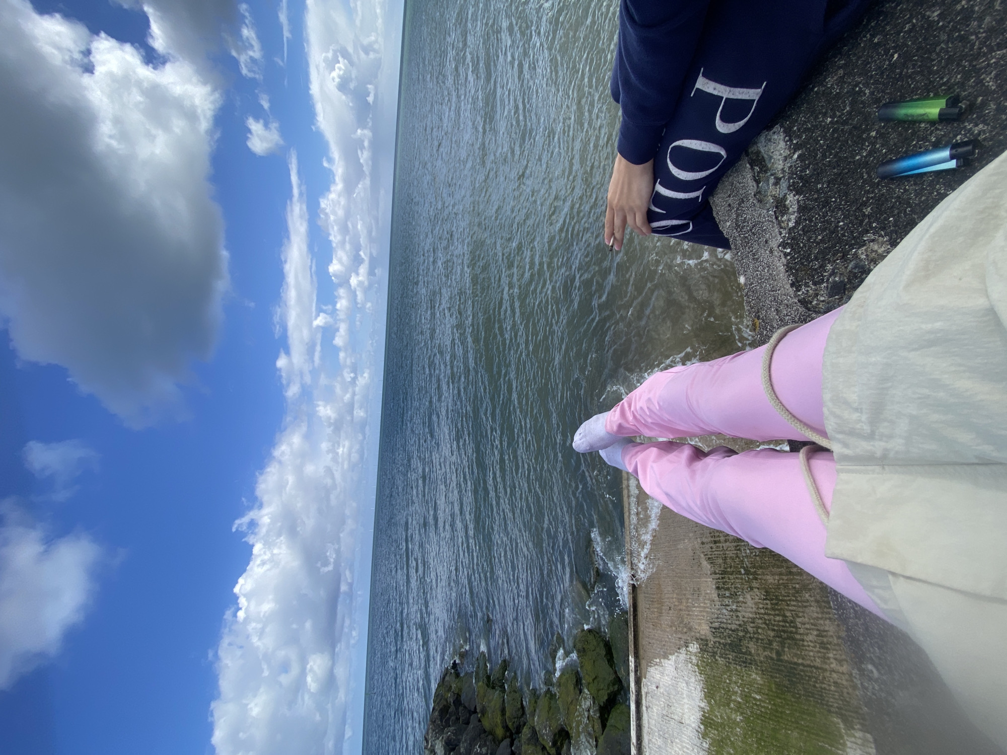 A medium shot captures two people sitting on a concrete structure overlooking a body of water. The camera is positioned to focus on their legs and the water. The person on the left wears dark blue Polo-branded pants and the person on the right wears pink pants with light colored socks and jacket. Two vape pens sit beside the person wearing the dark blue pants. The concrete structure where they sit appears to be a pier, with waves lapping against the edge. Below the pier is a rocky shoreline covered in green algae. The background consists of a wide expanse of water under a partially cloudy sky. The location is likely the beach at Old Colwyn in the United Kingdom.