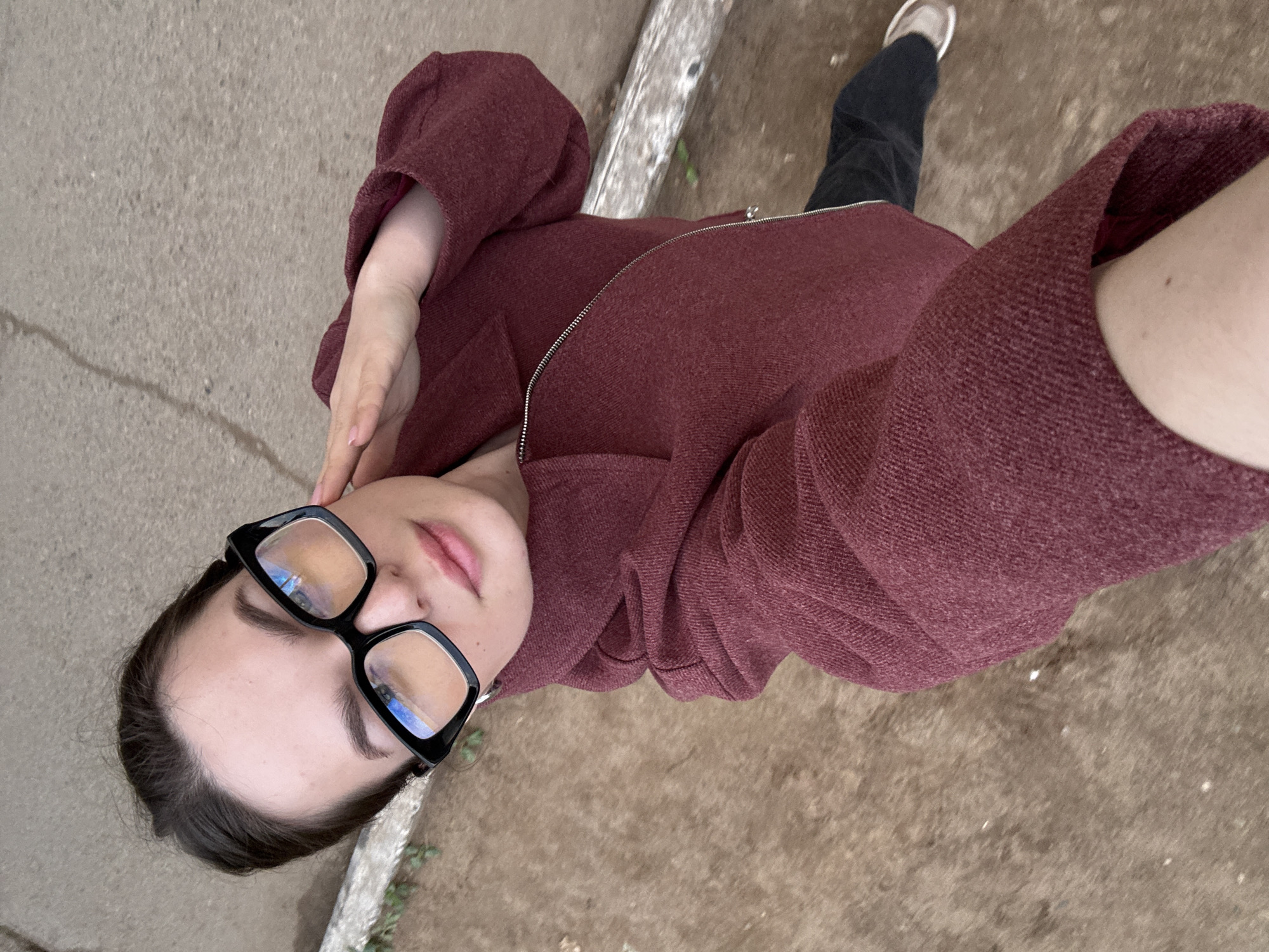 A young woman is taking a selfie in Chaikovsky, Russia. She is wearing black-framed glasses and a maroon zip-up jacket, with one hand raised to her forehead. She is standing on an unpaved surface with a concrete curb nearby. Her shoe and the lower part of her jeans are visible in the background.