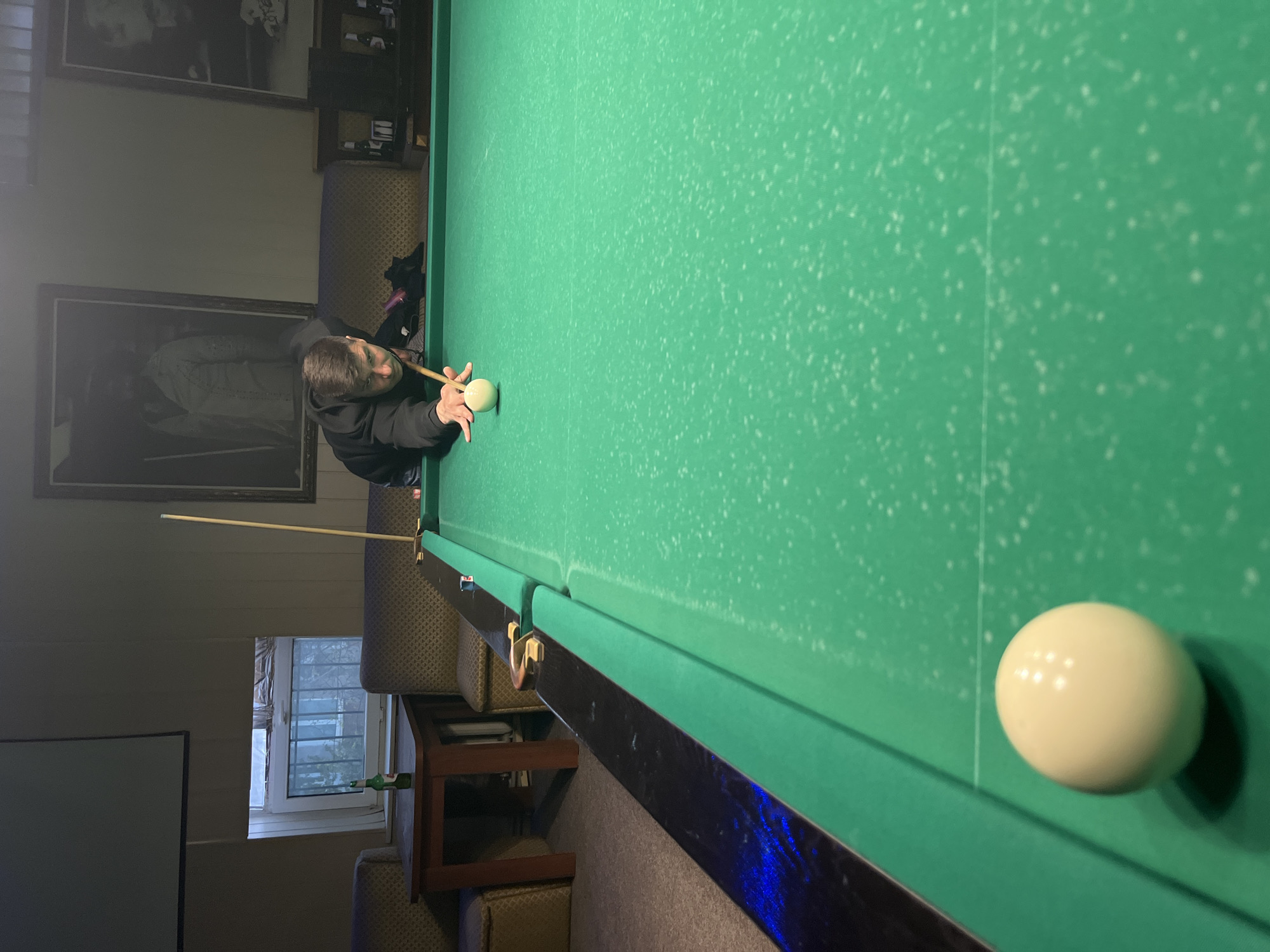 A person in a black hooded sweatshirt is seen playing billiards in a room setting. They are positioned at the edge of a green billiard table, holding a cue stick aimed at a cue ball that is also on the table. The table is in a room with framed pictures on the walls, and a couch behind the person. There is also a window with bars visible in the background. The location is in the city of Unknown, Unknown.