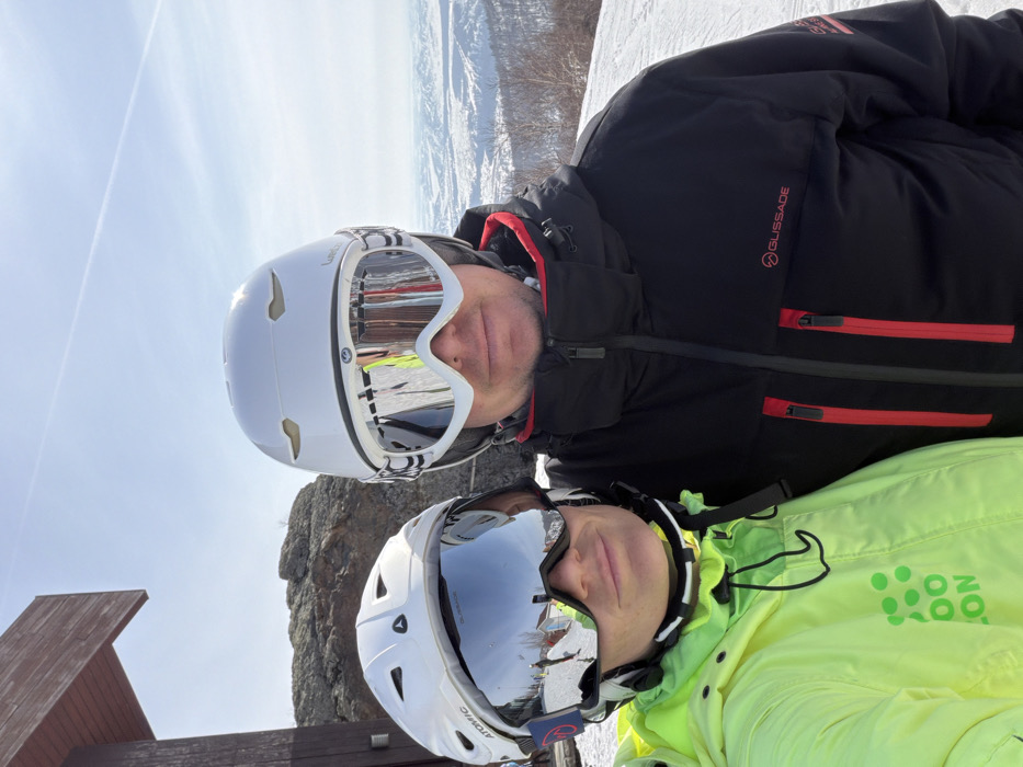 Two individuals, an adult male and an adult female, are depicted outdoors in a snowy, mountainous environment at a ski resort in Zelenaya Polyana, Russia. Both subjects are wearing white ski helmets and mirrored goggles. The female, positioned on the left, wears a neon yellow ski jacket with a partially visible green logo. Her white helmet is branded "ATOMIC." The male, positioned on the right, wears a black ski jacket featuring red zipper accents and a "GLISSADE" logo; his white helmet is branded "WED'ZE." They are in close proximity, facing the camera, likely posing for a selfie. The background displays snow-covered terrain, distant snow-capped hills, exposed rock formations, and a partial view of a dark brown wooden structure on the far left. The sky is clear blue with a visible contrail. The scene indicates a bright, sunny day on a ski slope.