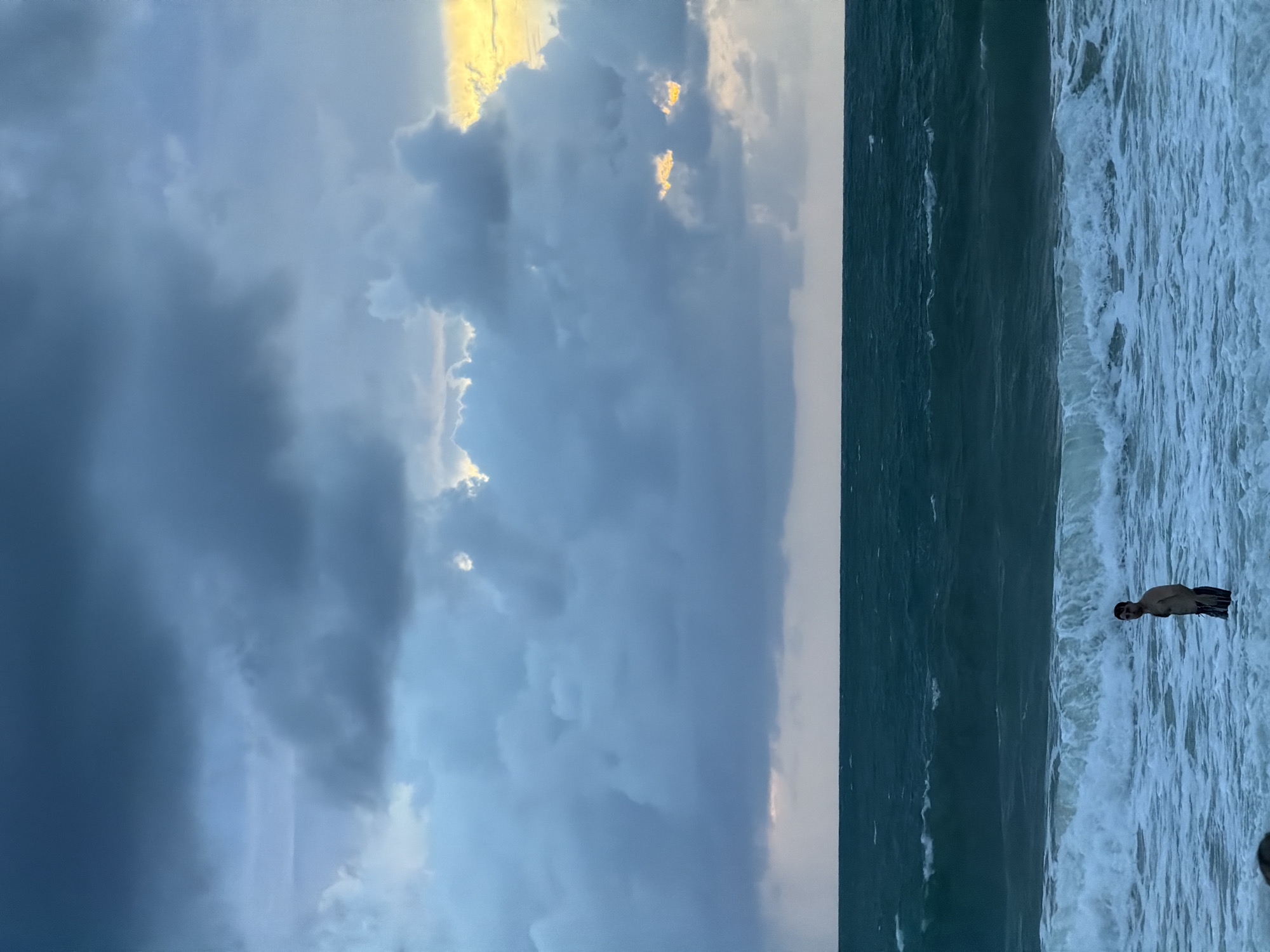 The image shows a coastal scene at Choeng Thale, Thailand. The sky is predominantly filled with storm clouds, with some areas illuminated by sunlight. The sea is turbulent, with waves crashing onto the shore. A lone male figure stands in the water, facing the waves.