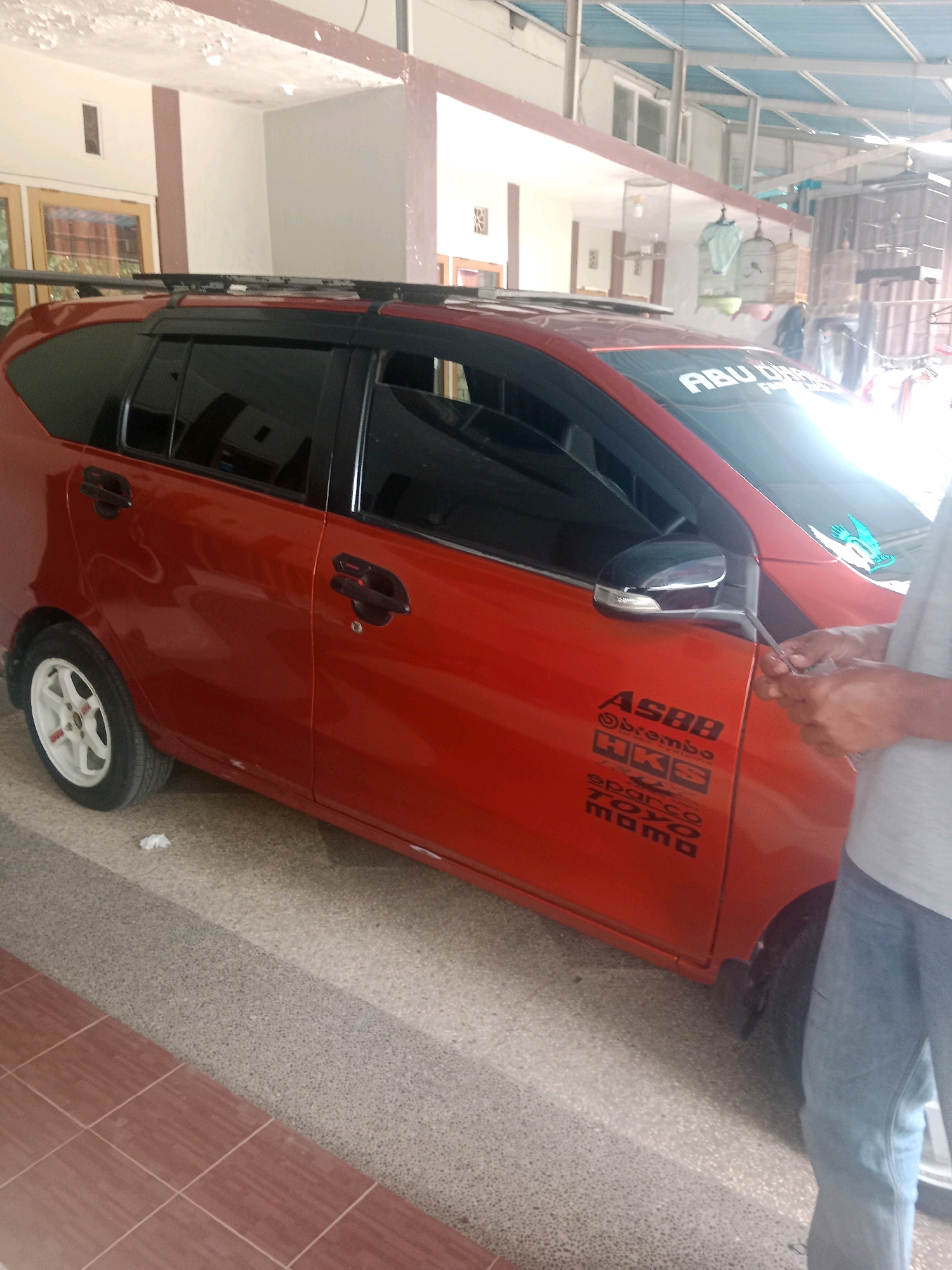 A red four-door car is parked on a speckled gray and red tile floor. The car has a black roof rack and tinted windows. Decals are affixed to the side of the car, including "ASBB," "Obrembo," "HKS," "sparco," "TOYO," and "mama." A person in blue jeans and a gray shirt stands to the right of the car, holding a small tool and looking at the car. In the background, there are buildings with white and brown walls, a blue metal roof structure, and several bird cages hanging from the ceiling. The scene appears to be in a covered outdoor area, possibly a carport or a workshop, in Banjarbaru, Indonesia, given the architectural style and the presence of bird cages.