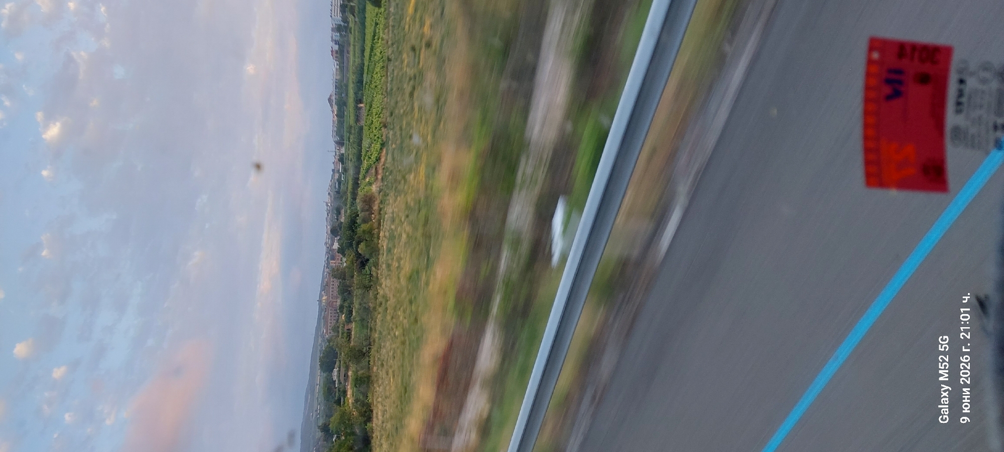 The image depicts a landscape scene viewed from a moving vehicle, likely a train or car, as evidenced by the motion blur. The foreground shows a grey asphalt road with a bright blue line marking the edge, and a silver metal guardrail. A red sticker with white lettering is visible on the right side of the road. In the middle ground, there is a blurred grassy field with some vegetation. The background features a panoramic view of a town or city with buildings and greenery, set against a sky with clouds. The date and time "9 June 2026, 21:01" and "Galaxy M52 5G" are overlaid in the bottom left corner.
The town depicted in the background is Vilafranca del Penedès, Spain. The prominent building visible in the mid-background, to the left of the center, is the Basilica of Santa Maria. The vineyards characteristic of the Penedès wine region are also visible.