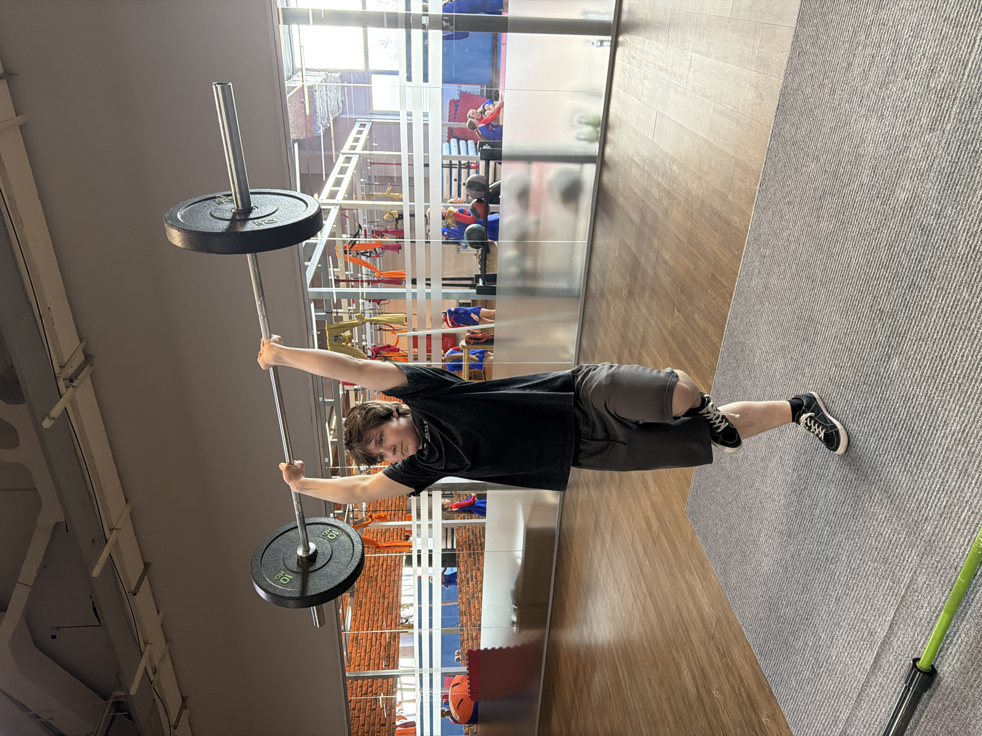 An indoor, horizontally oriented image, showing a young male actively engaged in weightlifting within a modern gym setting in Chaikovsky, Russia. The subject, positioned centrally, is holding a barbell with two 10 kg black weight plates overhead with both hands, arms extended. He wears a black short-sleeved t-shirt (with partial, illegible text, possibly "ALEJAH"), dark grey shorts, and black sneakers with white soles. His expression is focused, looking towards the camera.
The gym features a wooden laminate floor on the left and a textured grey carpet or matting on the right. A tall, reflective glass partition runs horizontally through the middle ground, showcasing reflections of other gym areas and individuals, suggesting other activities are taking place. In the background, behind the partition, various gym equipment such as suspension trainers, resistance bands, medicine balls, and possibly pull-up bars are visible. The gym walls appear to be a mix of brick and possibly painted surfaces, with large windows allowing natural light, indicating it is daytime. A green object, possibly part of a jump rope handle, is visible on the grey carpet in the foreground.