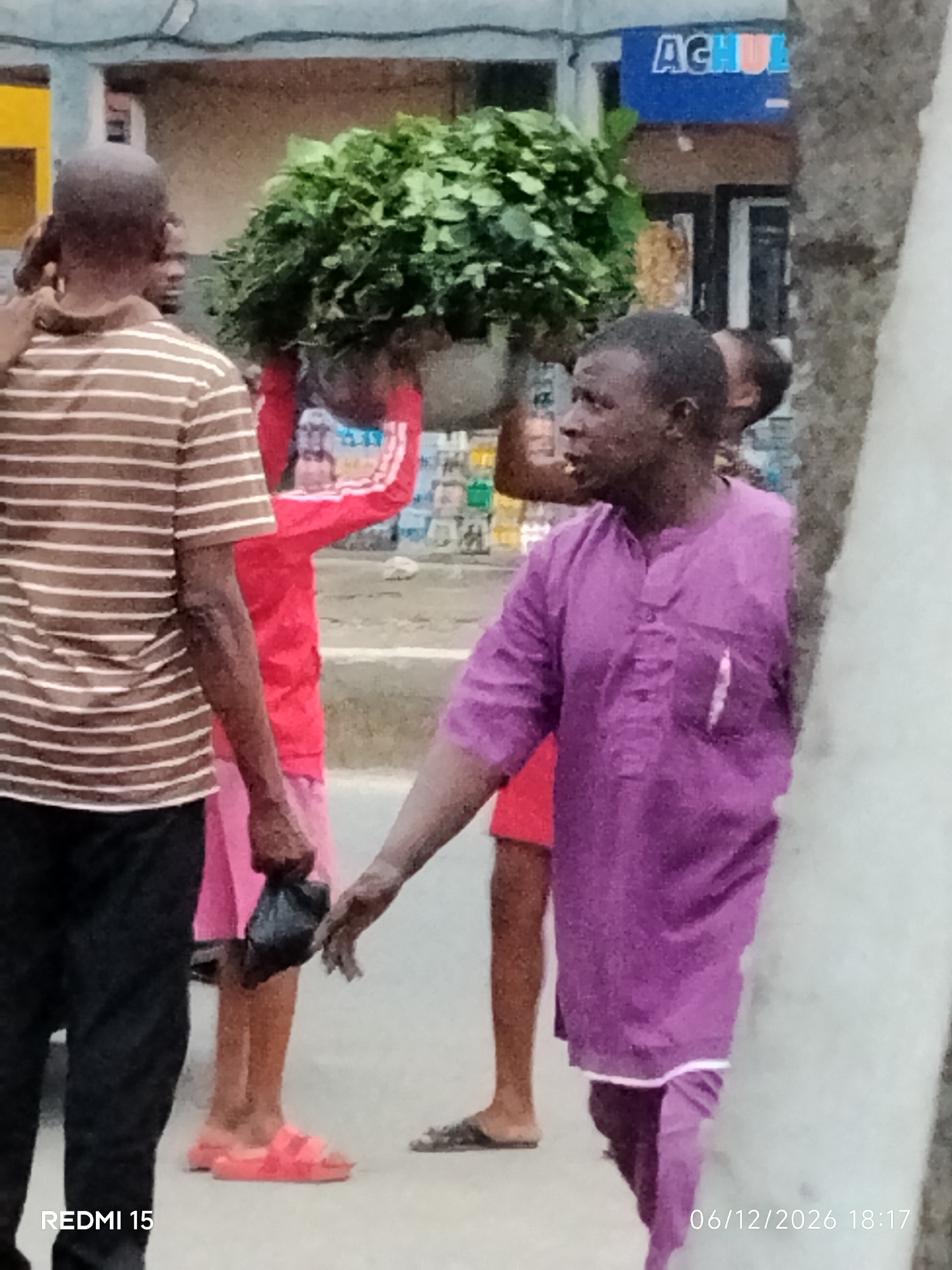 The image depicts a street scene in Aba, Nigeria, with several individuals visible. In the foreground, a man wearing a brown and white striped t-shirt and black pants stands facing away from the camera, his left arm obscured. To his right, a man in a purple traditional outfit is the primary subject. He is looking to his right, with his mouth slightly open. Behind him, a person in a red jacket and pink shorts is carrying a large bundle of green vegetation on their head. Further in the background, several other individuals and buildings are visible, including a blue sign with white lettering that appears to say "AGHUA". The environment suggests an outdoor marketplace or busy street. The lighting is diffused, indicating it might be daytime. A timestamp at the bottom right corner reads "06/12/2026 18:17".