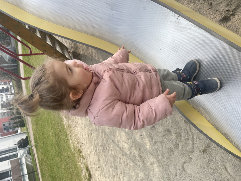 The scene depicts a playground in the city of Senden, Germany, under daylight conditions. A single toddler, estimated to be between 18 months and 3 years old, is positioned on a metal playground slide. The child has light brown hair tied in a high bun, wears small stud earrings, and is dressed in a pink hooded puffer jacket, light grey pants, and dark blue winter boots with colorful motifs. The child is standing upright on the textured surface of the slide, which has yellow side rails, and is looking towards the viewer's left with an open mouth. Their right arm is extended, with the index finger pointing downwards towards the slide's surface. The ground immediately surrounding the slide consists of sandy material. In the background, a grassy area is visible to the left, beyond which modern multi-story residential buildings with balconies are present. To the right of the child, a wooden structure with red metal rails, likely part of the slide's access stairs, is partially visible.