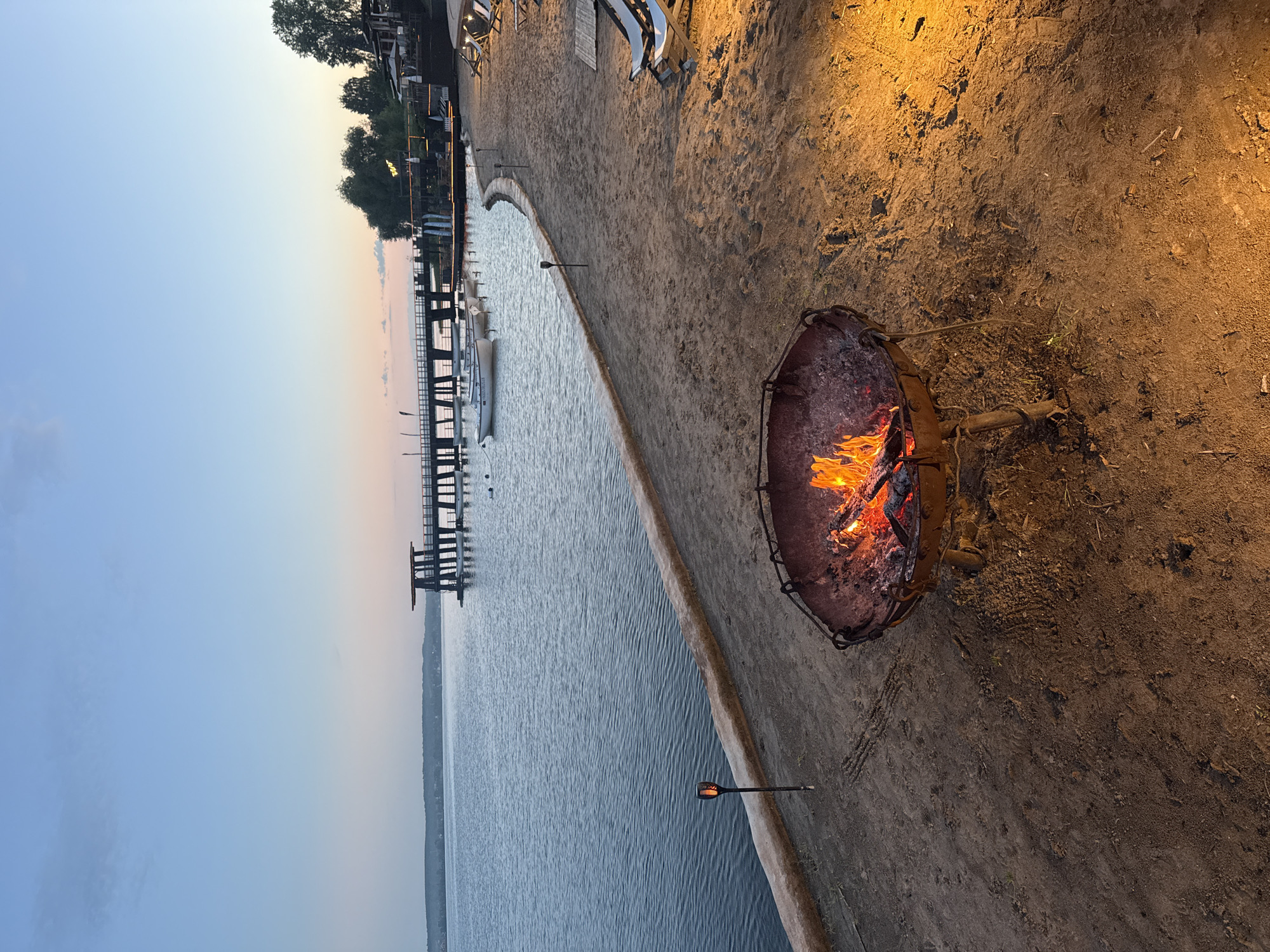 This image depicts a tranquil waterfront scene at twilight, likely situated on the Votkinsk Reservoir in the city of Votkinsk, Russia, at a recreational complex such as "Якорь" (Anchor) or "Пляж "Водники"" (Vodniki Beach). The sky, occupying the upper third, displays a clear gradient from light blue at the zenith to a soft, pale orange-pink on the horizon, indicative of either dusk or dawn.
The foreground features a sandy or dirt beach area. Dominating the immediate left foreground is a rusty, metal fire pit containing actively burning wood and glowing embers, producing visible flames and casting warm orange light onto the surrounding ground. The ground immediately around the fire pit is uneven, consisting of packed earth with some sparse plant growth and faint tracks. To the right, a strong, localized artificial light source casts an intense orange glow across the sand. Further right and extending into the mid-ground, several reclined beach chairs with white fabric are arranged on the sand, accompanied by a light-colored mat. Along the curved edge where the beach meets the water, several slender, dark-colored torch-like lights with illuminated, flame-simulating tops are spaced.
The mid-ground is dominated by a calm, dark blue body of water, showing minimal ripples. A prominent, dark wooden pier extends from the right into the water, featuring a distinctive elevated structure at its end with a dark roof and multiple supporting columns. Several small, white rowboats are moored near the pier, some displaying faint text such as "HOUSE OF MUSIC". On the distant shore, a line of dark green trees is visible, silhouetted against the horizon, with additional illuminated structures and trees located behind the pier complex on the right side of the image. No people or animals are present in the scene.