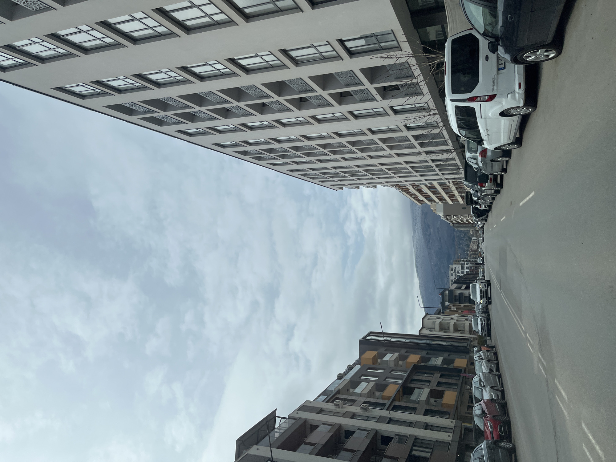 The image shows a street scene in Sofia, Bulgaria. The view is down a street lined with modern apartment buildings on both sides. Cars are parked along the right side of the street. In the distance, a mountain range is visible under a cloudy sky. The buildings have multiple stories, with windows and balconies. The street is paved and has painted lane markers. There are no people visible in the shot.