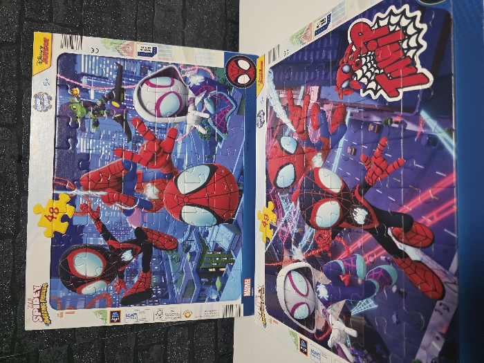 The image displays two partially assembled jigsaw puzzles, stacked one above the other, both featuring characters from "Marvel's Spidey and His Amazing Friends." The items are situated against a dark, textured background resembling a brick wall on the left.
The top puzzle, predominantly visible, depicts four animated characters within a stylized cityscape at dusk. The characters include Spider-Man (classic red and blue suit), Miles Morales (black and red suit), Ghost-Spider (white and purple suit), and Green Goblin (green suit with a purple glider). Spider-Man is positioned centrally, reaching outwards. Miles Morales is to the left, and Ghost-Spider is to the right. Green Goblin is in the upper right, flying on his glider. The cityscape background features various illuminated buildings. A yellow puzzle piece bearing the number "48" is placed in the upper left quadrant. Branding elements visible include "Marvel Spidey and His Amazing Friends," "Glow in the dark," "5+," and "Disney Junior." The puzzle is incomplete, with several individual pieces visible.
The bottom puzzle is mostly assembled and positioned beneath the top one, displaying a different cityscape scene, primarily at night with prominent neon pink and blue light trails. This puzzle features Miles Morales in the foreground, facing right, with Ghost-Spider to his left, and classic Spider-Man to his right. All characters are in dynamic, action-oriented poses. A yellow puzzle piece with the number "48" is in the upper left, and a graphic of a "Thwip!" speech bubble is in the lower right corner. The packaging suggests a 48-piece count for both puzzles.
There are no discernible visual cues or text within the image that link the depicted scenes or the objects themselves to a specific location within the city of Zweibrücken, Germany. The backgrounds are generic, animated cityscapes.