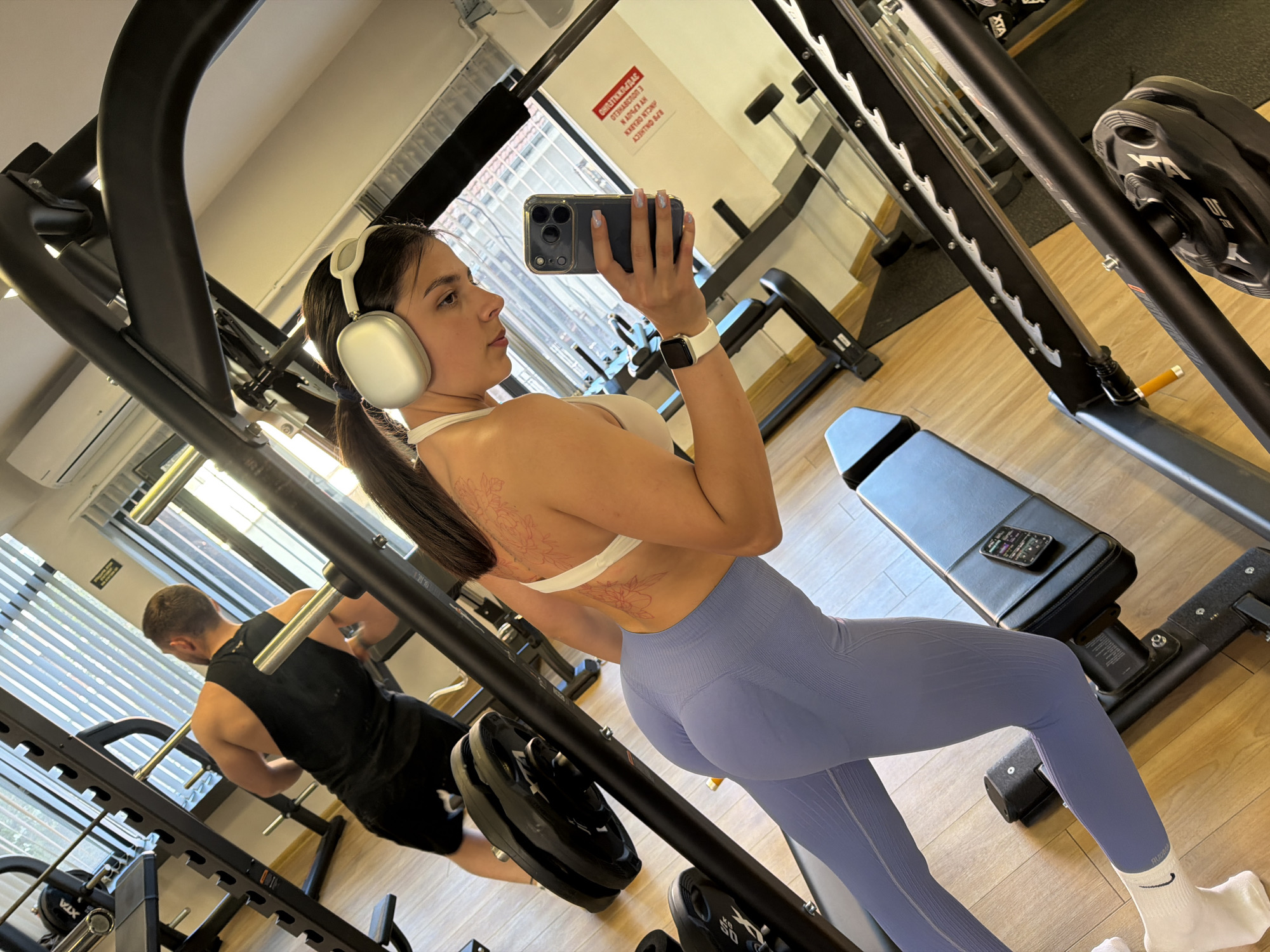 The image depicts a woman in a gym, likely located in Gabrovo, Bulgaria. She is holding a smartphone in her right hand, presumably taking a selfie, while wearing a white sports bra, light blue leggings, and white headphones. She has a tattoo on her upper back. A man in a black shirt and shorts is visible in the background, engaging in a workout. Gym equipment, including weight racks and benches, are also present. The gym has a wooden floor and appears to be well-lit. A sign with text in Bulgarian is visible on the wall in the background.