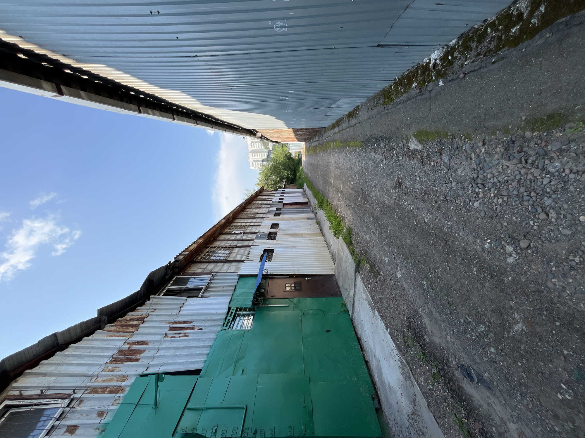 A narrow, unpaved alleyway between two corrugated metal buildings in Krasnoyarsk, Russia, under a clear blue sky with scattered clouds. The building on the left is a mix of faded white corrugated metal with rust stains and a large, bright green metal structure covering part of its facade. It has several windows, some with protective bars. A brown door is visible on this building, with a small blue awning above it. The building on the right is entirely clad in grey corrugated metal. The alleyway is covered in gravel and dirt, with some patches of dampness and greenery. In the distance, the alley opens up to a view of trees and a multi-story apartment building. The scene depicts a utilitarian and somewhat weathered urban environment.