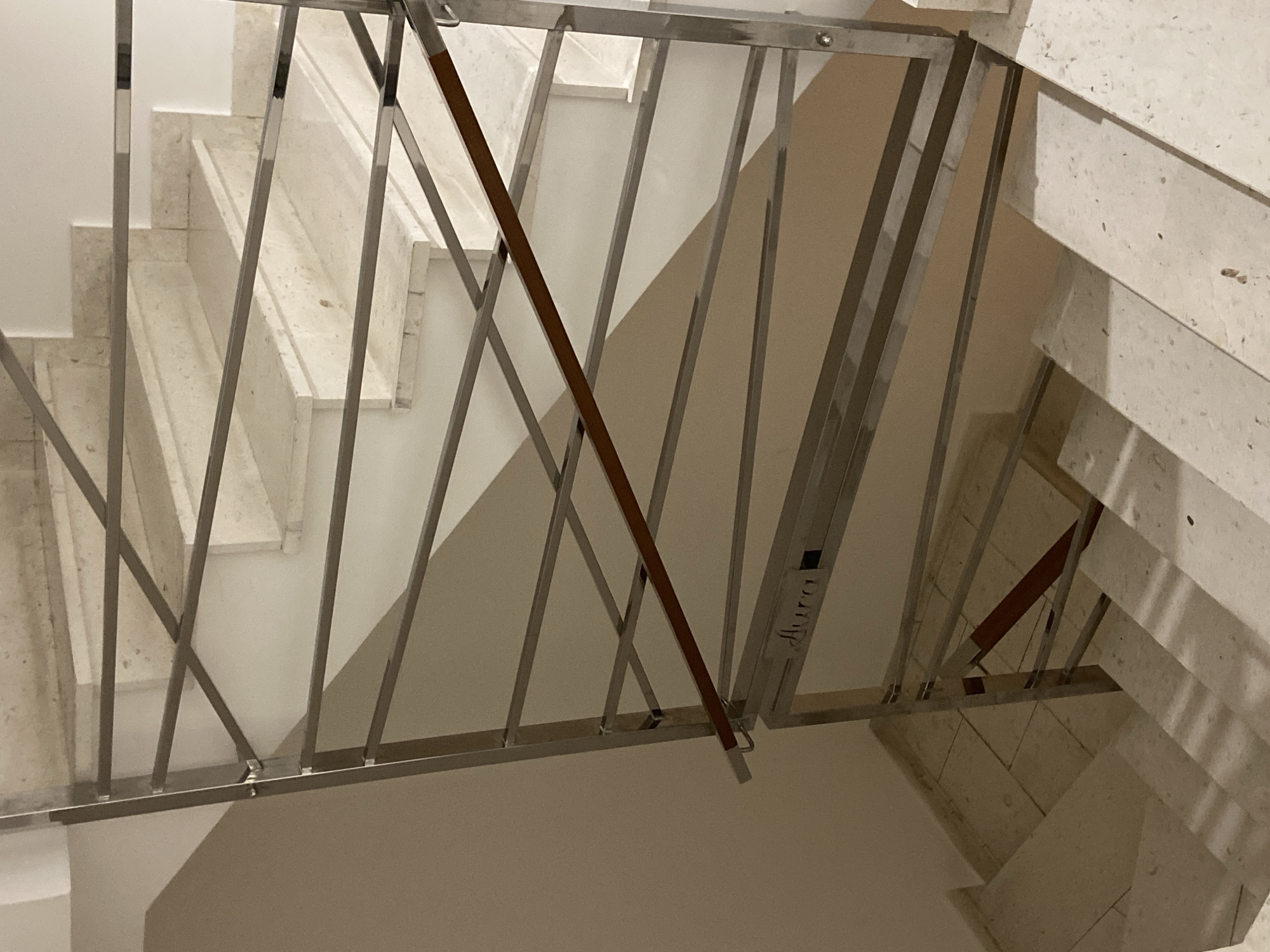This image depicts an indoor stairwell within a building in Budva, Montenegro. The multi-level staircase is constructed from light-colored, speckled marble or stone. A modern balustrade, made of polished metal with horizontal bars, runs adjacent to the staircase. A diagonal wooden handrail is integrated into the upper section of the metal balustrade. A sign with the word "Aura" is attached to the metal balustrade at a lower landing. The walls are painted a light, neutral color, displaying distinct shadows. The scene is illuminated by artificial indoor lighting. No individuals or active subjects are present.