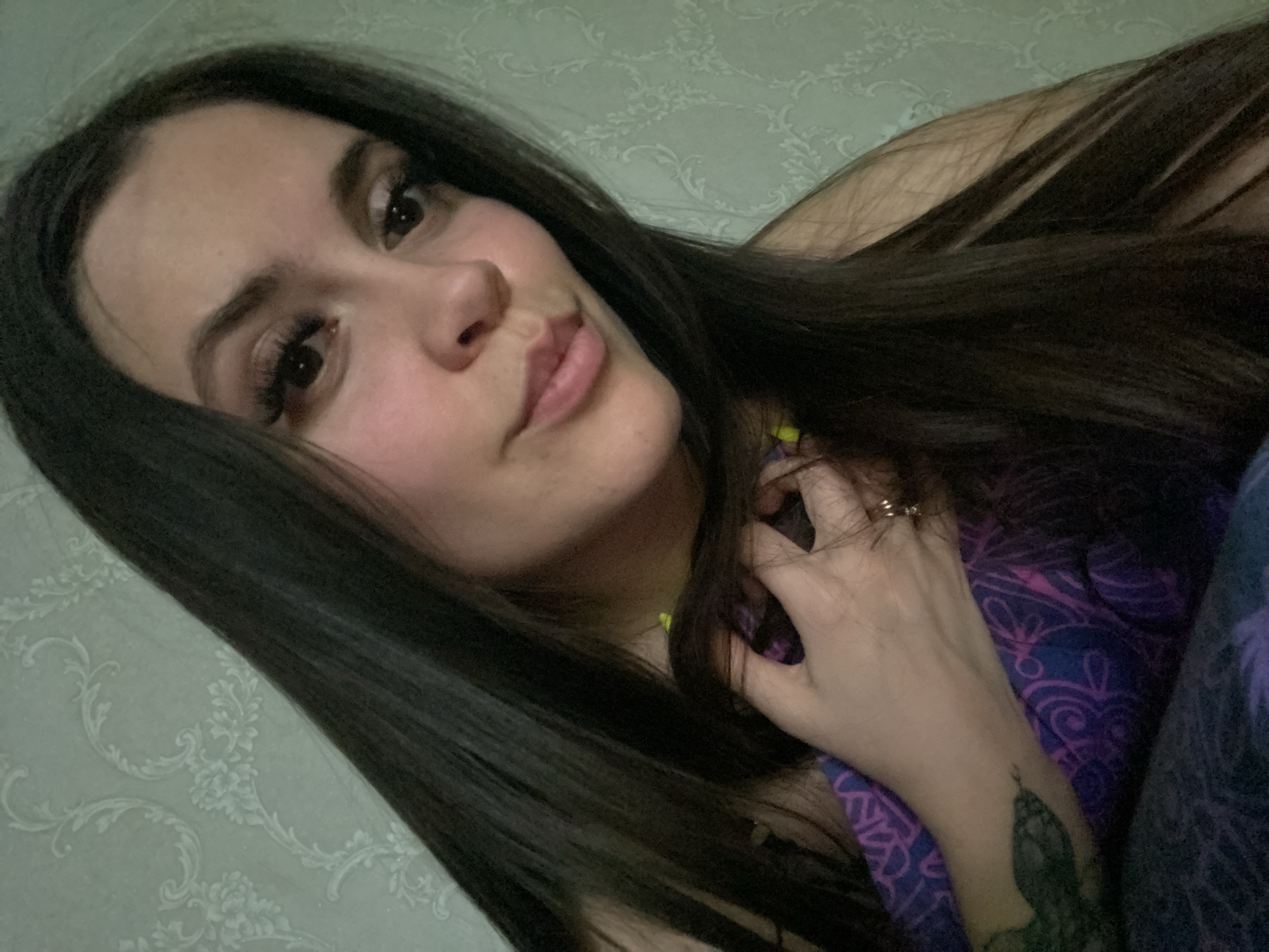 A woman with long, dark hair is pictured in a close-up selfie. She has light skin, dark eyes, and full lips. Her right hand, adorned with a ring, is near her face, partially obscuring the side of her neck. A glimpse of a purple and blue patterned garment is visible. A tattoo is present on her left forearm. The background is a light green wall with a patterned design. The location is within Chaikovsky, Russia, as stated in the prompt.