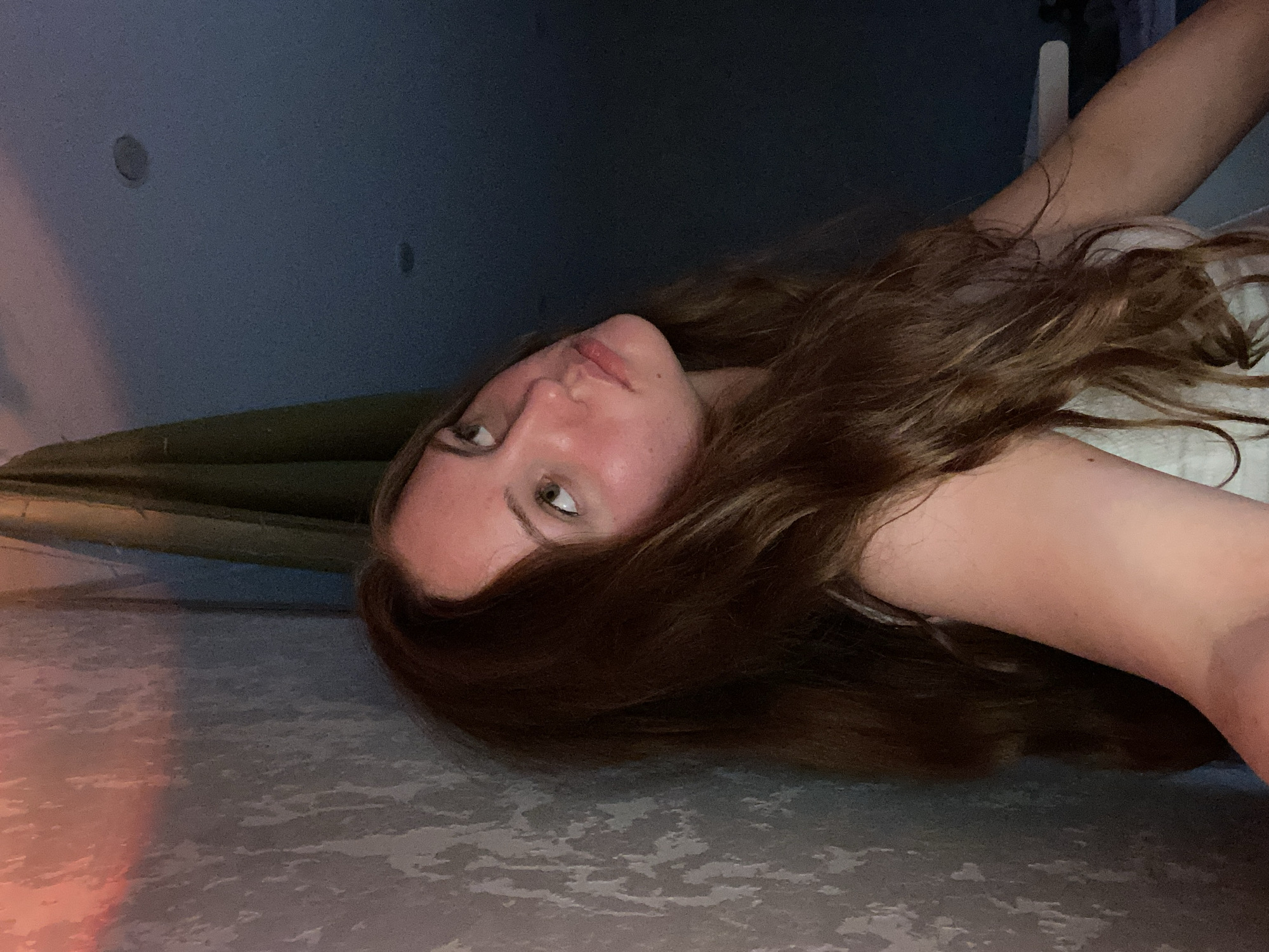 This candid photo, taken in Izhevsk, Russia, captures a young woman lying down in an indoor setting. She has long, wavy, reddish-brown hair spread around her head and fair skin with a natural blush on her cheeks. Her light-colored eyes are directed slightly upwards and off-camera, conveying a calm, thoughtful, or relaxed expression. She appears to be wearing a light-colored top, possibly white or off-white.
The background is dimly lit, suggesting evening or nighttime. In the upper left, a dark blue-gray wall is visible, featuring two small, circular, recessed objects, which could be lights or speakers. Below the wall, a dark green fabric, possibly part of a blanket or furniture, spans the mid-ground. The lower portion of the image shows a textured, light gray surface, likely a carpet or patterned floor, which reflects a soft reddish-orange light from the bottom left corner, adding to the ambient glow. There is no visible text in the image.