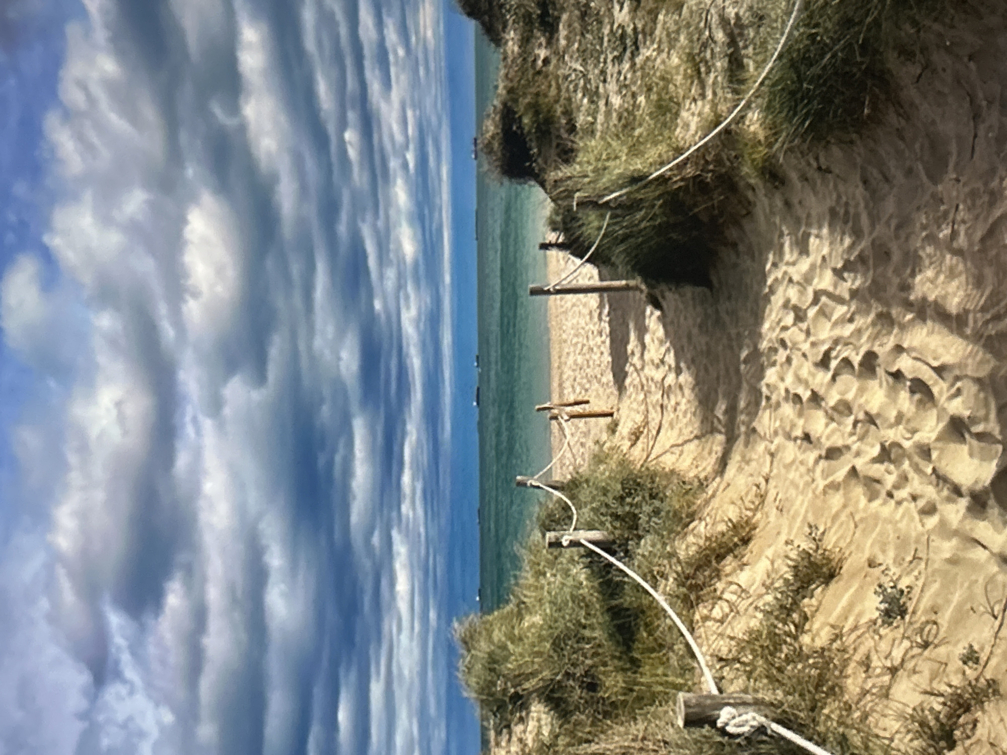 The image depicts a serene coastal landscape, viewed from a slightly elevated perspective looking towards the sea. A sandy path, illuminated by sunlight, winds down from the right foreground towards the beach. Along the left edge of this path, a simple white rope fence, supported by several rustic wooden posts, delineates the route through the sandy terrain. Patches of green and brown vegetation, resembling grasses and low shrubs, grow on the sandy banks and dunes flanking the path.
In the midground, the path opens onto a tranquil beach of light-colored sand, which meets the clear, calm turquoise-blue water of the sea. The horizon line is visible across the center of the image, where the sea meets a partially cloudy sky. Several small, dark objects, possibly boats or markers, are faintly discernible on the distant water. The sky, which occupies the upper portion of the frame, features an expanse of blue interspersed with numerous elongated, horizontal clouds in various shades of white and grey, suggesting a bright yet partly overcast day. The lighting indicates it is daytime. There are no people visible, and no text is present in the image. The scene evokes a sense of peaceful natural beauty and an inviting route to the shore.