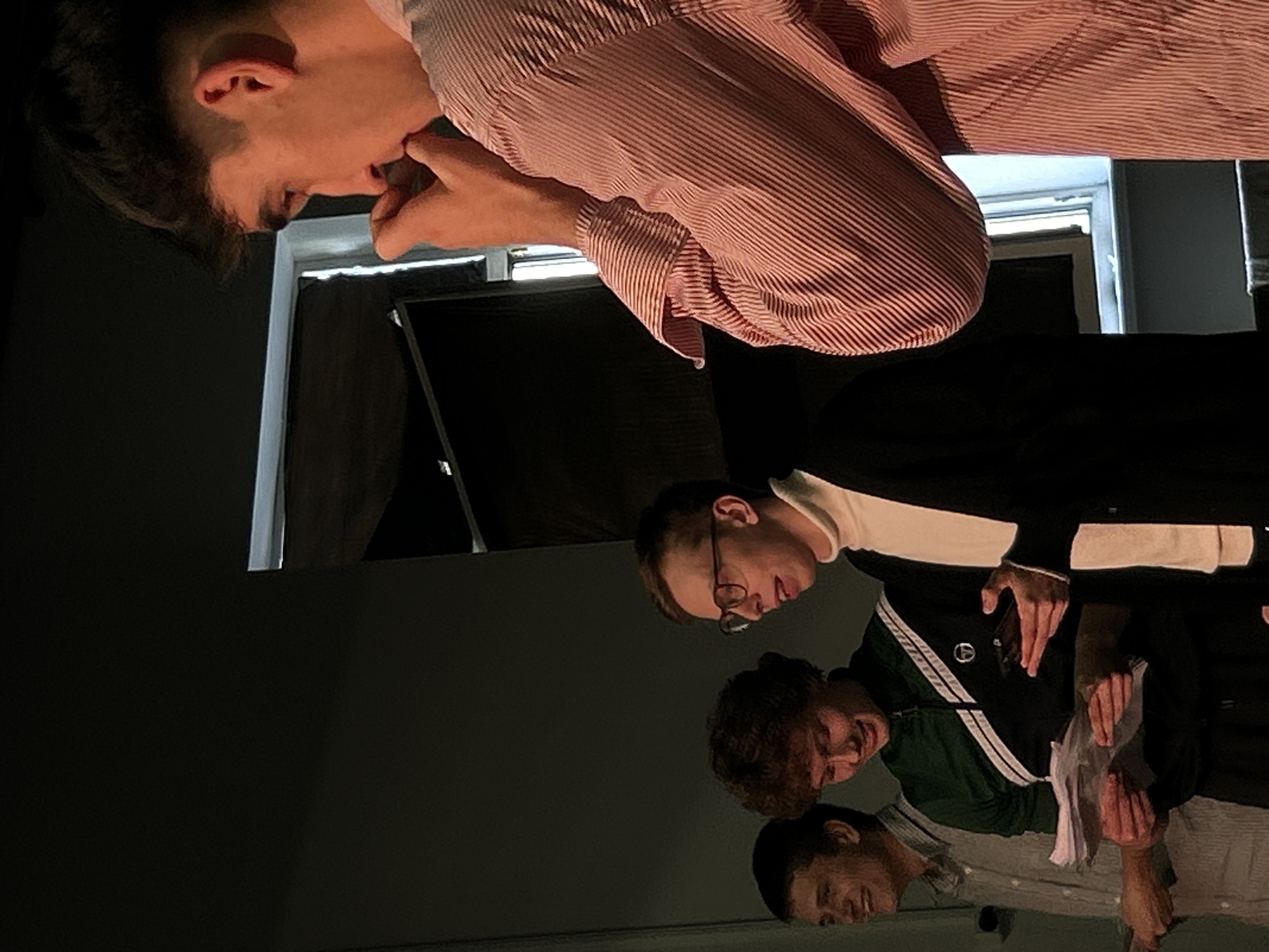 Four young men are standing in front of a dark wall, with a window visible in the background. The man on the far right is in profile, wearing a pink and white striped shirt and has his hand near his mouth. The man next to him is wearing glasses, a turtleneck, and a black cardigan, and is holding a phone. The man in the green jacket is holding papers, as is the man to his left who is wearing a grey shirt. The lighting is dim, with a spotlight effect on the men. The scene appears to be an indoor event or presentation.
